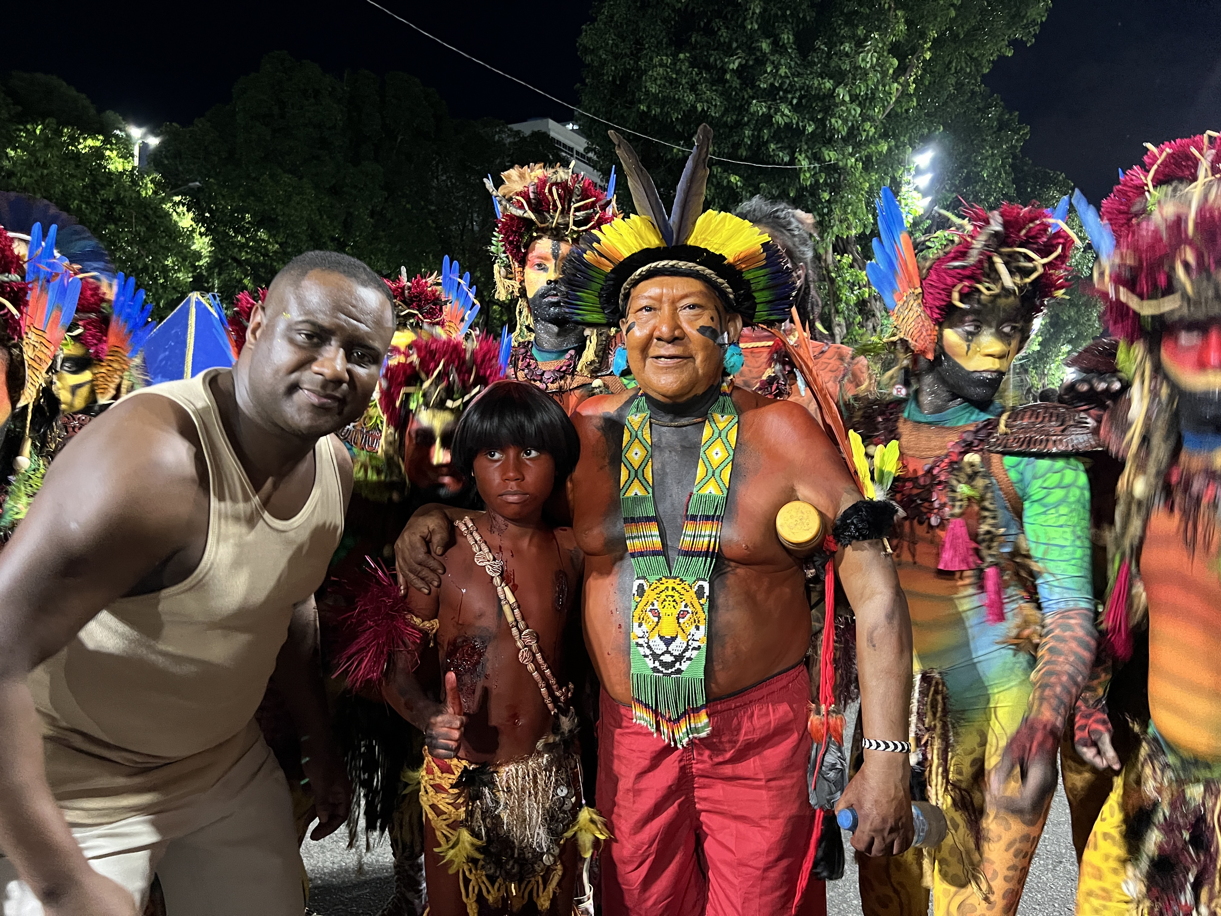 Davi Kopenawa poses for a photo with parade participants at Rio de Janeiro's Sambadrome. He wears a radiating feather headdress, red shorts and a beaded necklace.