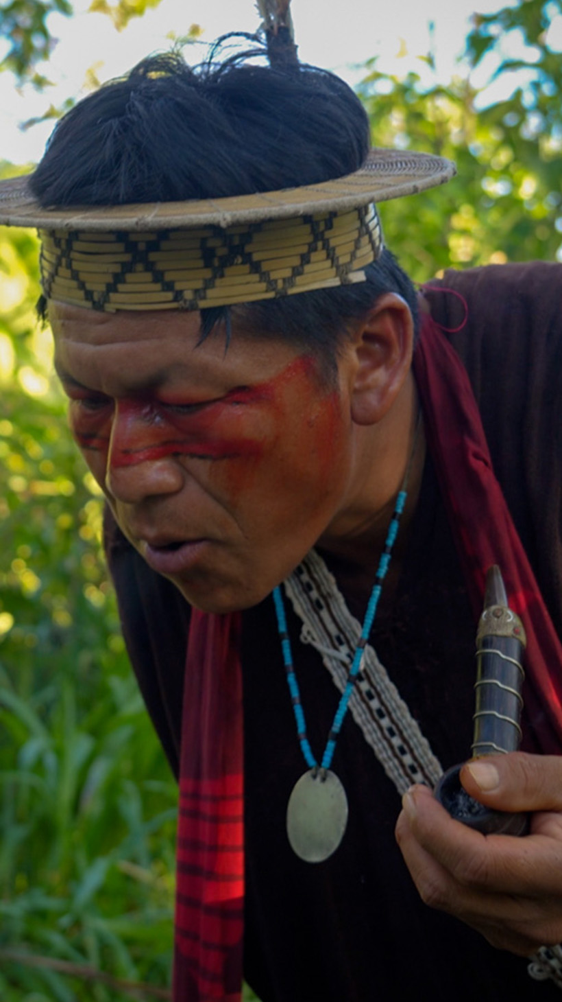 Ashaninka leader Tsitsiri Samaniego — dressed in a tunic and a straw headdress with three macaw feathers sticking upwards — inhales from a tobacco pipe as he stands before a headstone for his grandfather.