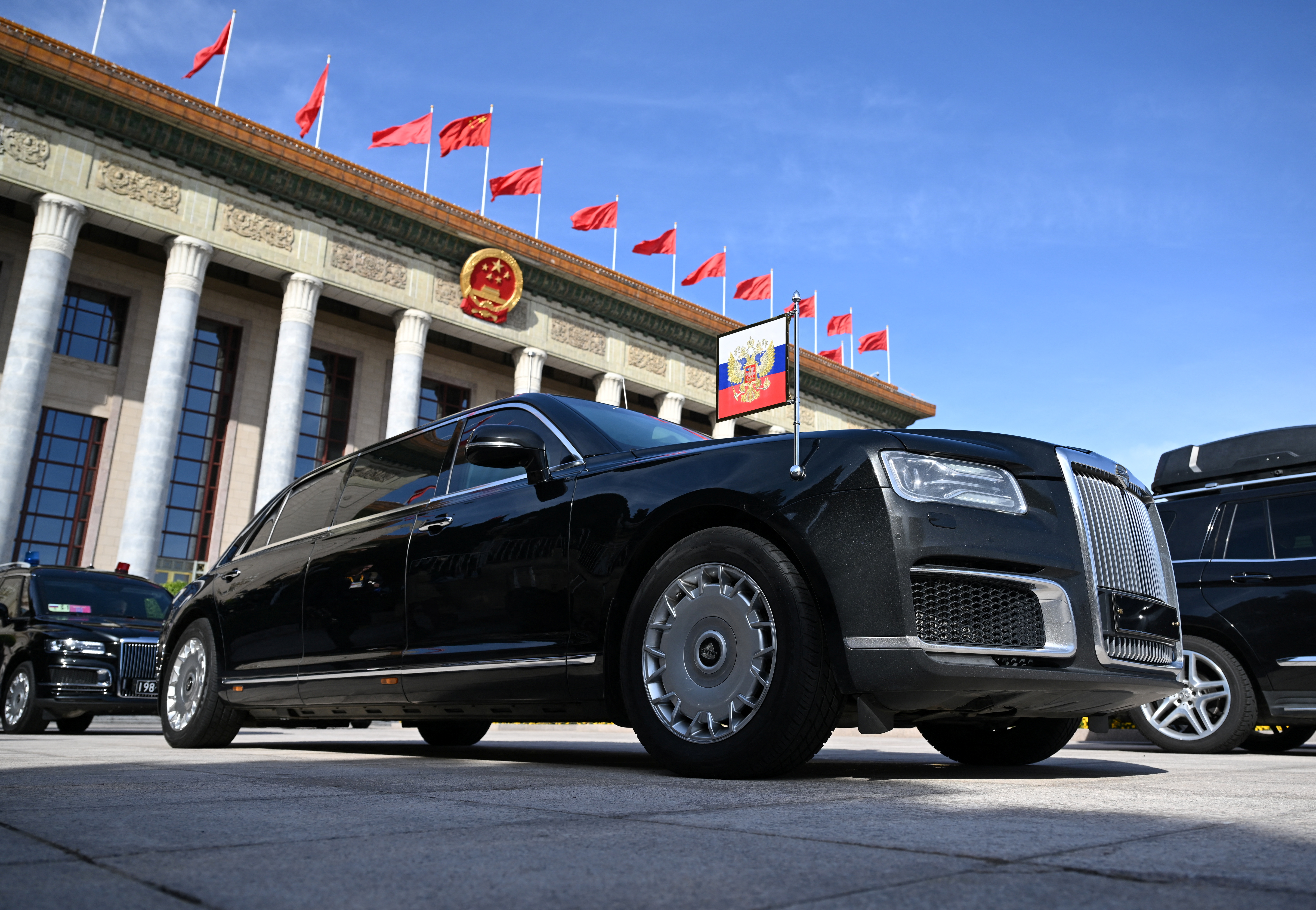 Vladimir Putin's Aurus outside the Great Hall of the Poeple in Beijing. There is a presidential pennant on the car's front right wing. The vehicle is black with a large grille at the front.