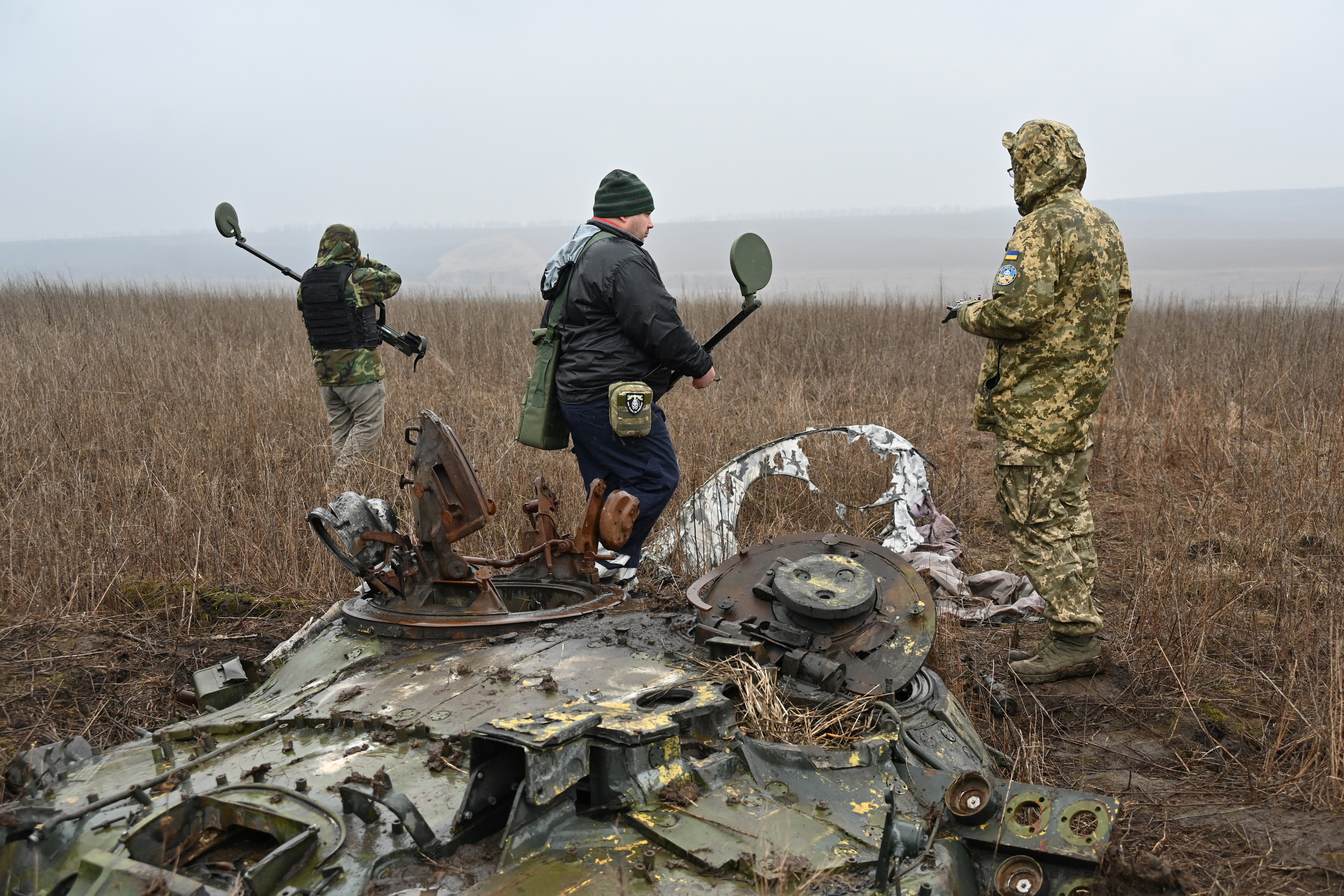 Deminers in Ukraine. There are three men holding metal detectors. They are in a field of high, brown grass.