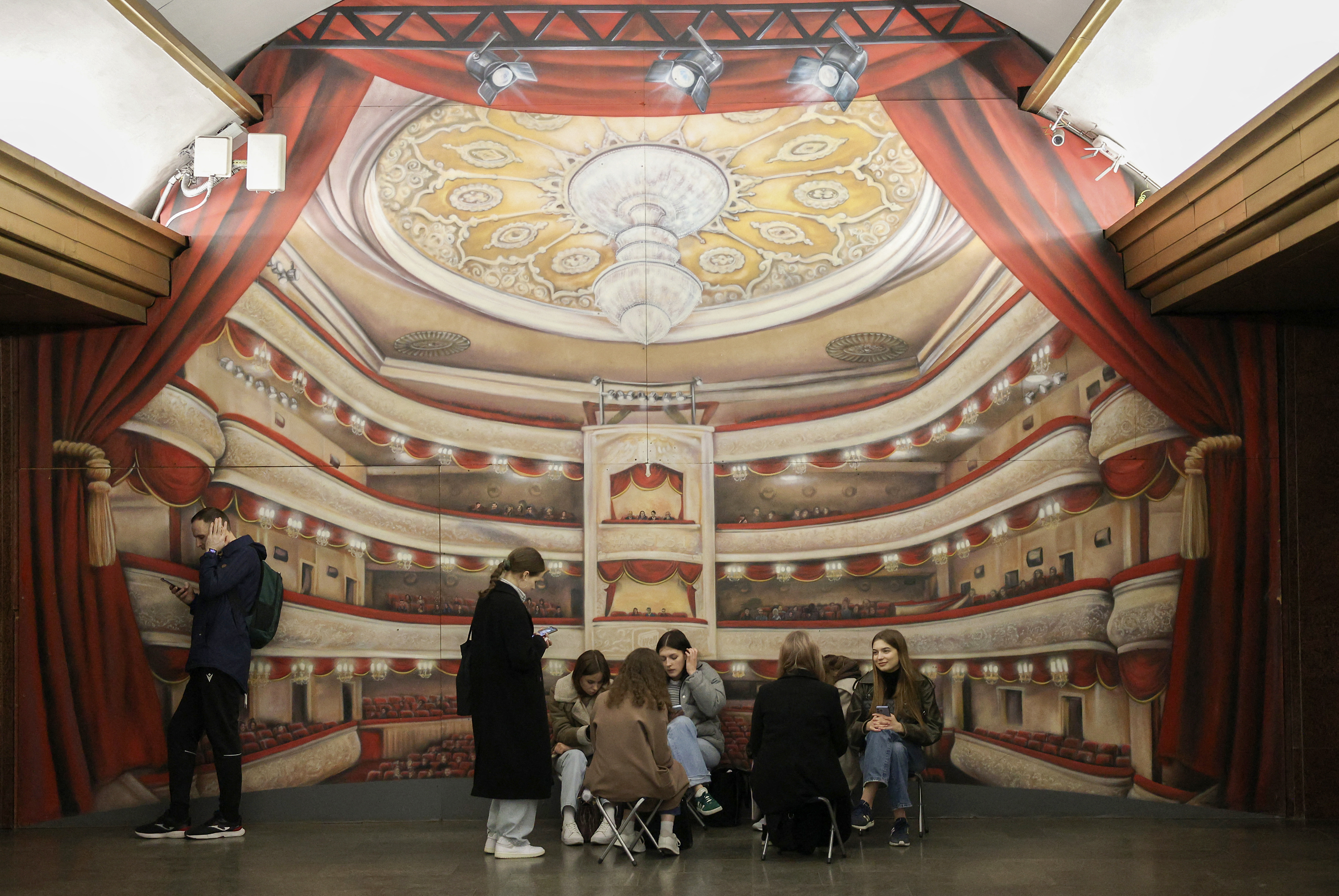 People taking shelter from an air raid in a Kyiv underground metro station The walls have been painted to look like a theatre auditorium.