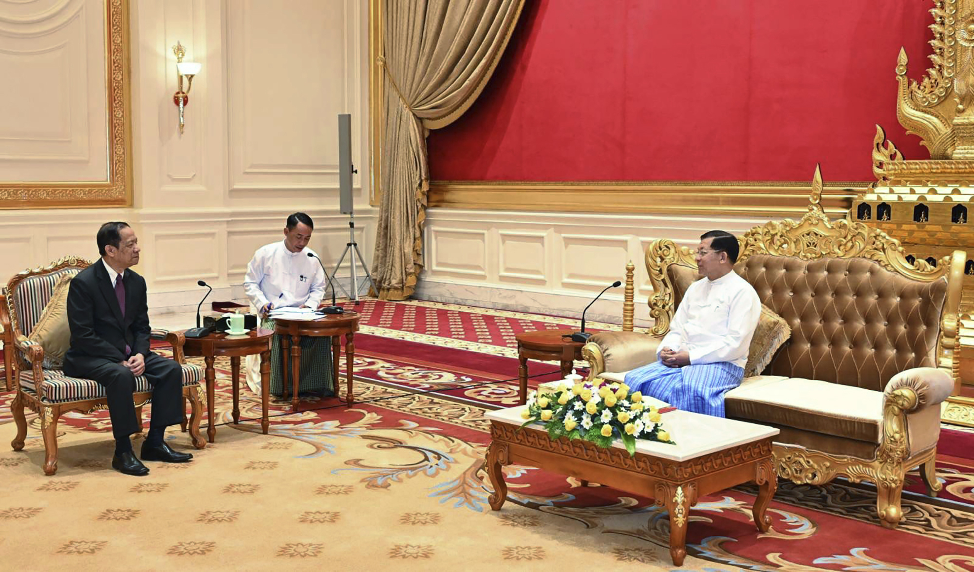 Laos appointed ASEAN special envoy Alounkeo Kittikhoun meets Min Aung Hlaing in Naypyidaw. Min Aung Hlaing is wearing a traditional Burmese outfit and is sitting on a large ornate sofa trimmed in gold. Alounkeo Kittikhoun is sitting at a distance to the general's right in an upright chair. There is a desk with a large flower display in front of Min Aung Hlaing