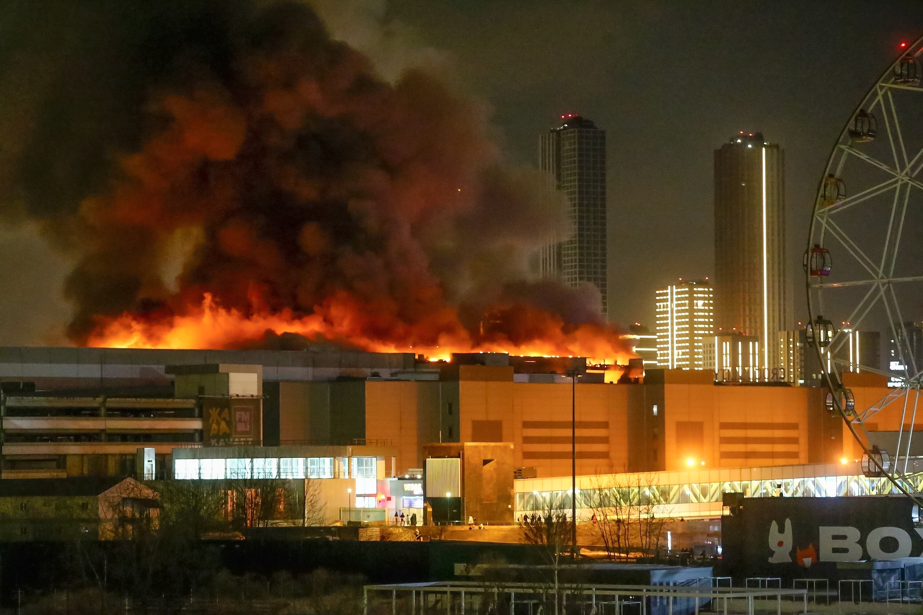 Crocus City Hall engulfed in fire. Orange flames and clouds of black smoke are rising from the roof. The area on the ground is lit up with fire engines. 