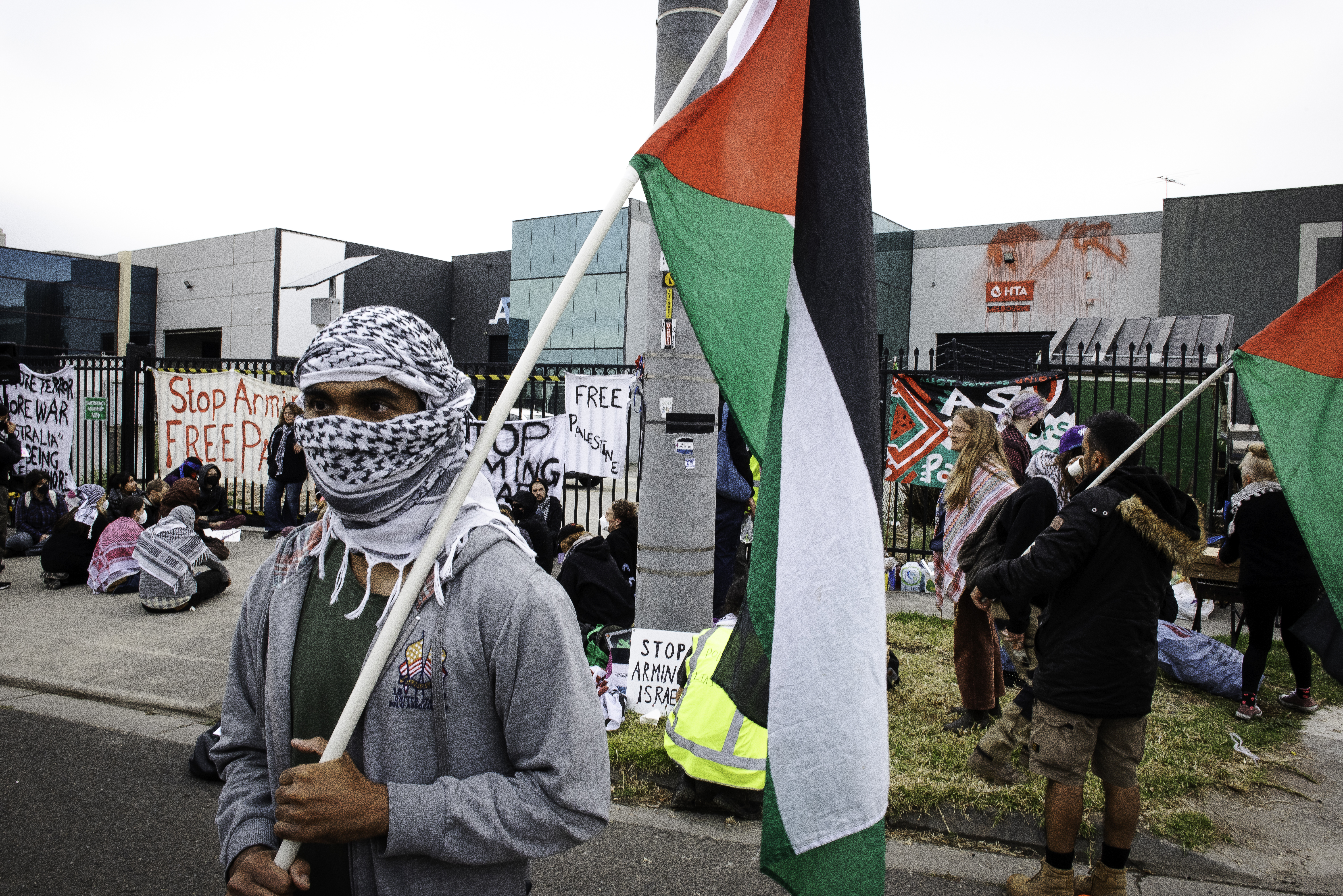 A protester carrying a Palestinian flag at a picket outside an Australian arms company. They have wrapped a Palesinian scarf around their face so only their eyes are visible, Other protesters are behind them. They have placards. Some are sitting on the ground.