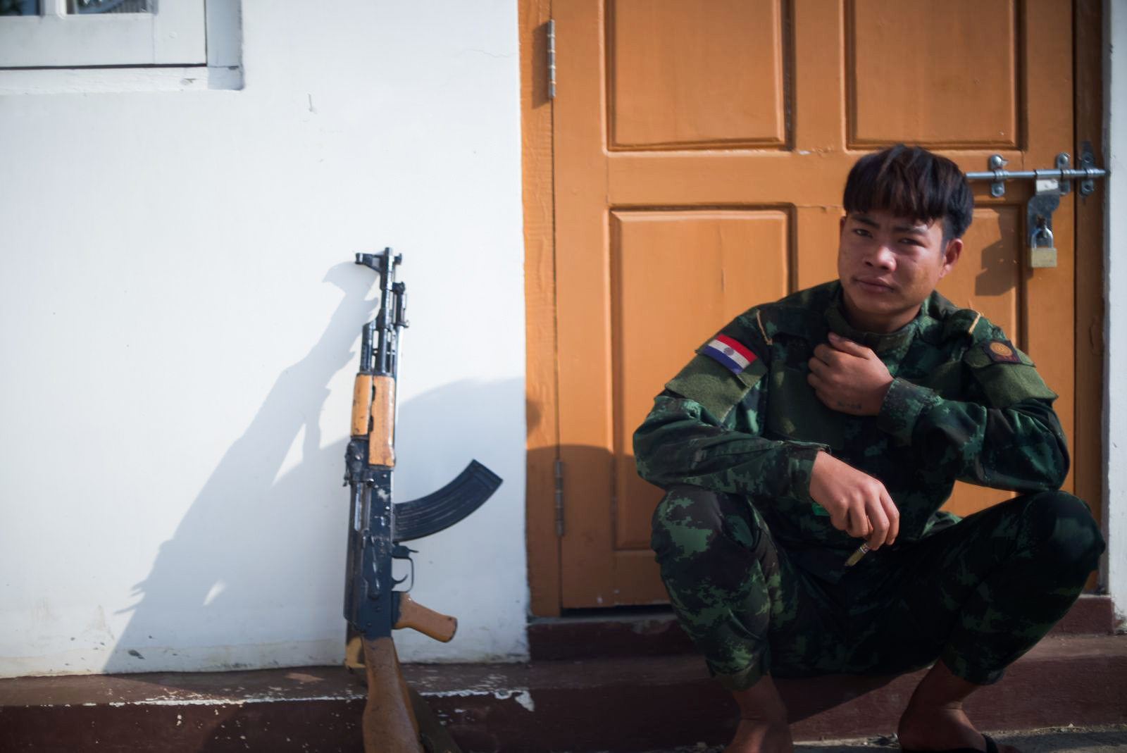 A fighter in Kayah. He is seated on the floor against a door. His weapon is propped against the wall next to him.