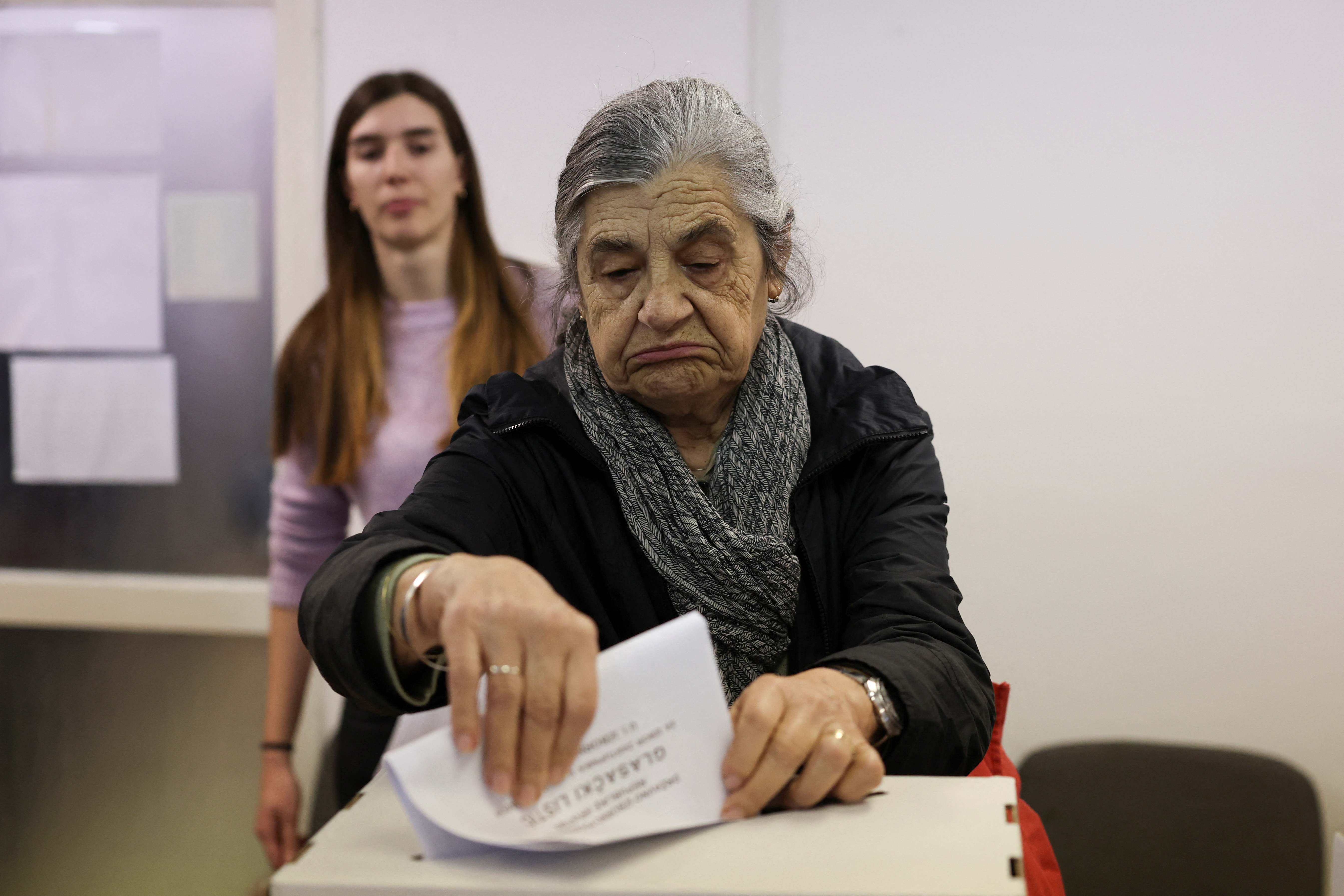 An older woman with grey hair putting her ballot paper in a ballot box. A younger woman is in the background.