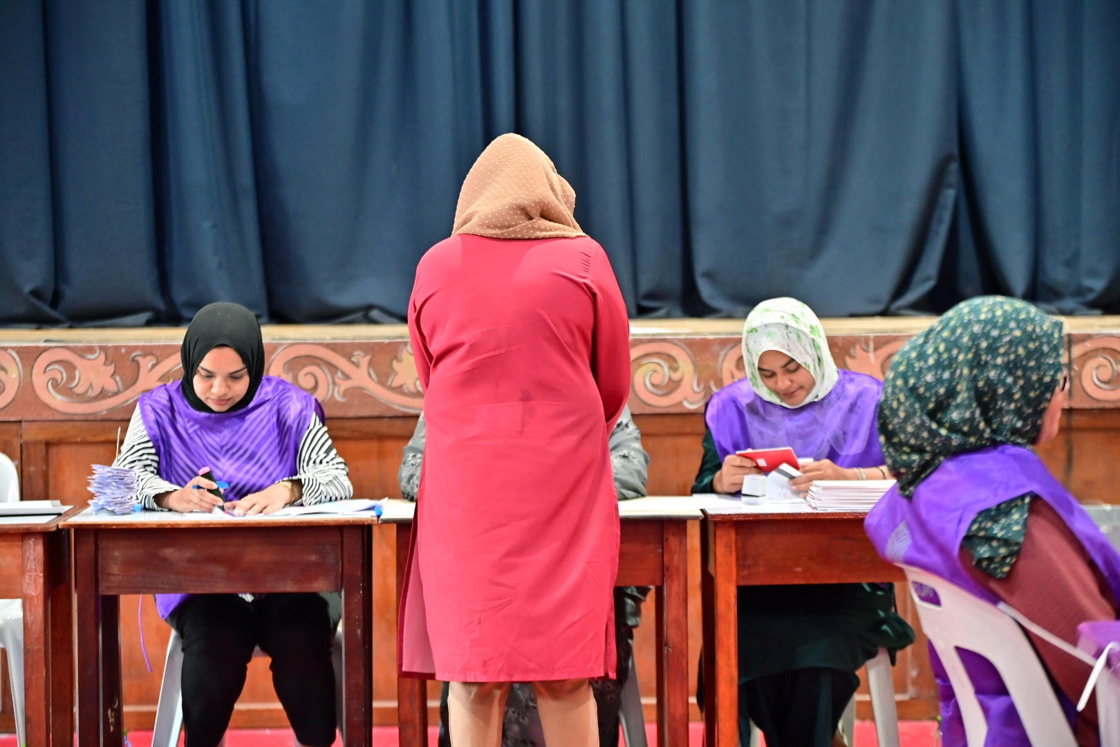 A voter casting her ballot in Madlives. She is wearing red and has her back to the camera. Elections officials in purple are facing her.