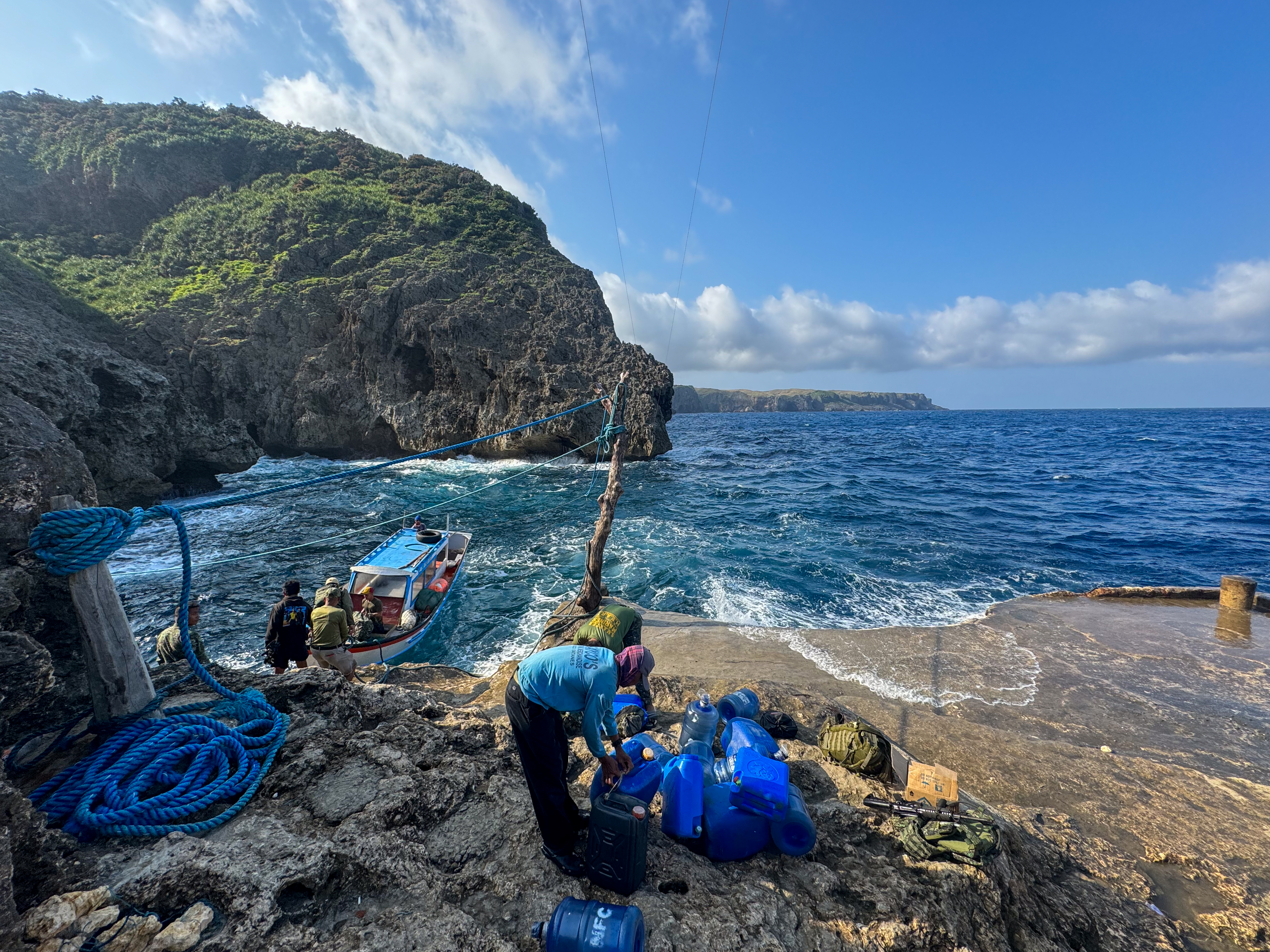A view of the port in Itbayat. There are jagged cliffs and rocks. A small boat is tied to the shore with ropes, People are waiting to board. The sea looks choppy. The island spreads out behind.