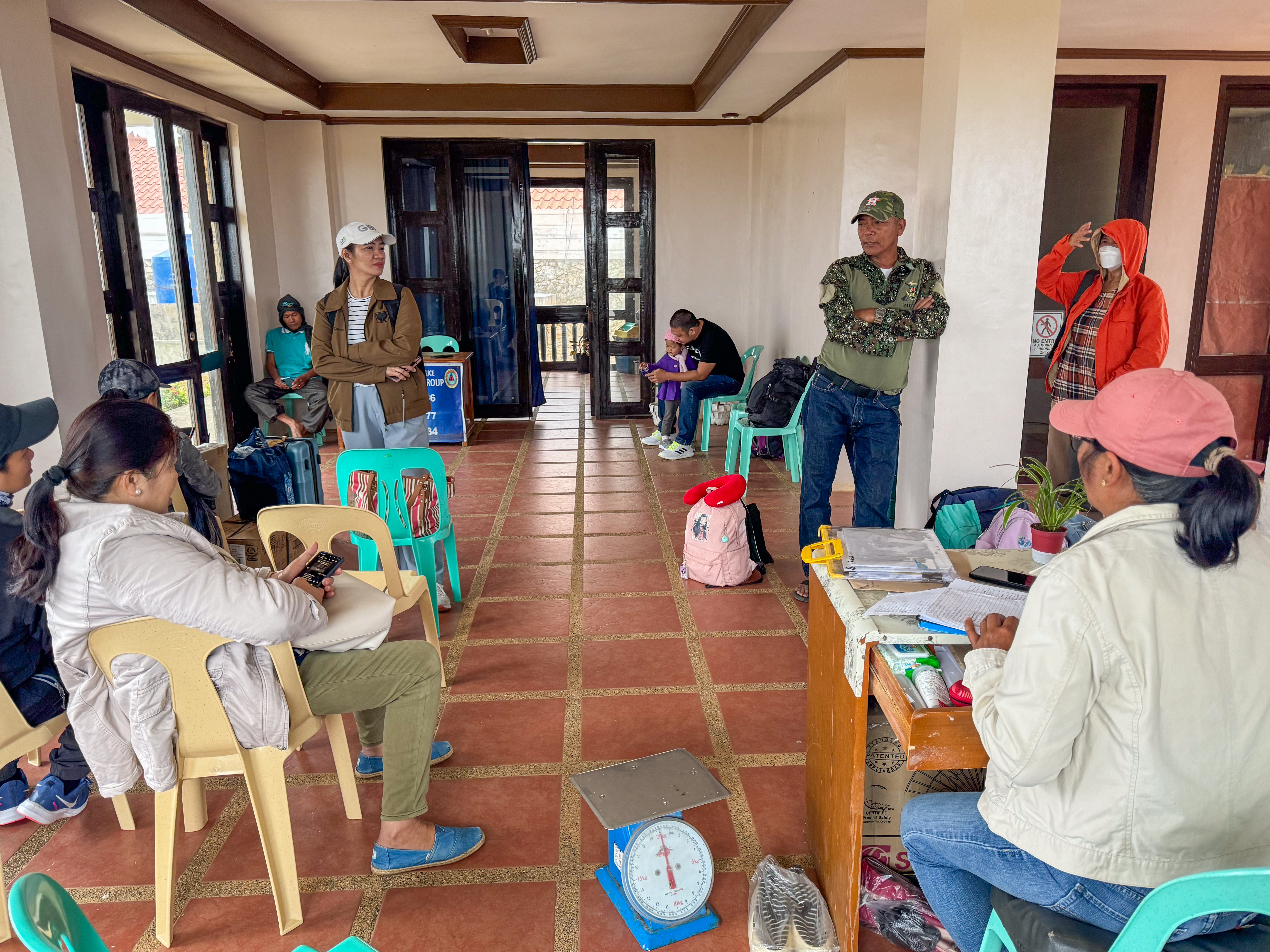 People in Itbayat's small airport discussing the call out for reservists. Some are standing. Some are seated on plastic chairs. There is a woman at a desk with lots of papers on it.