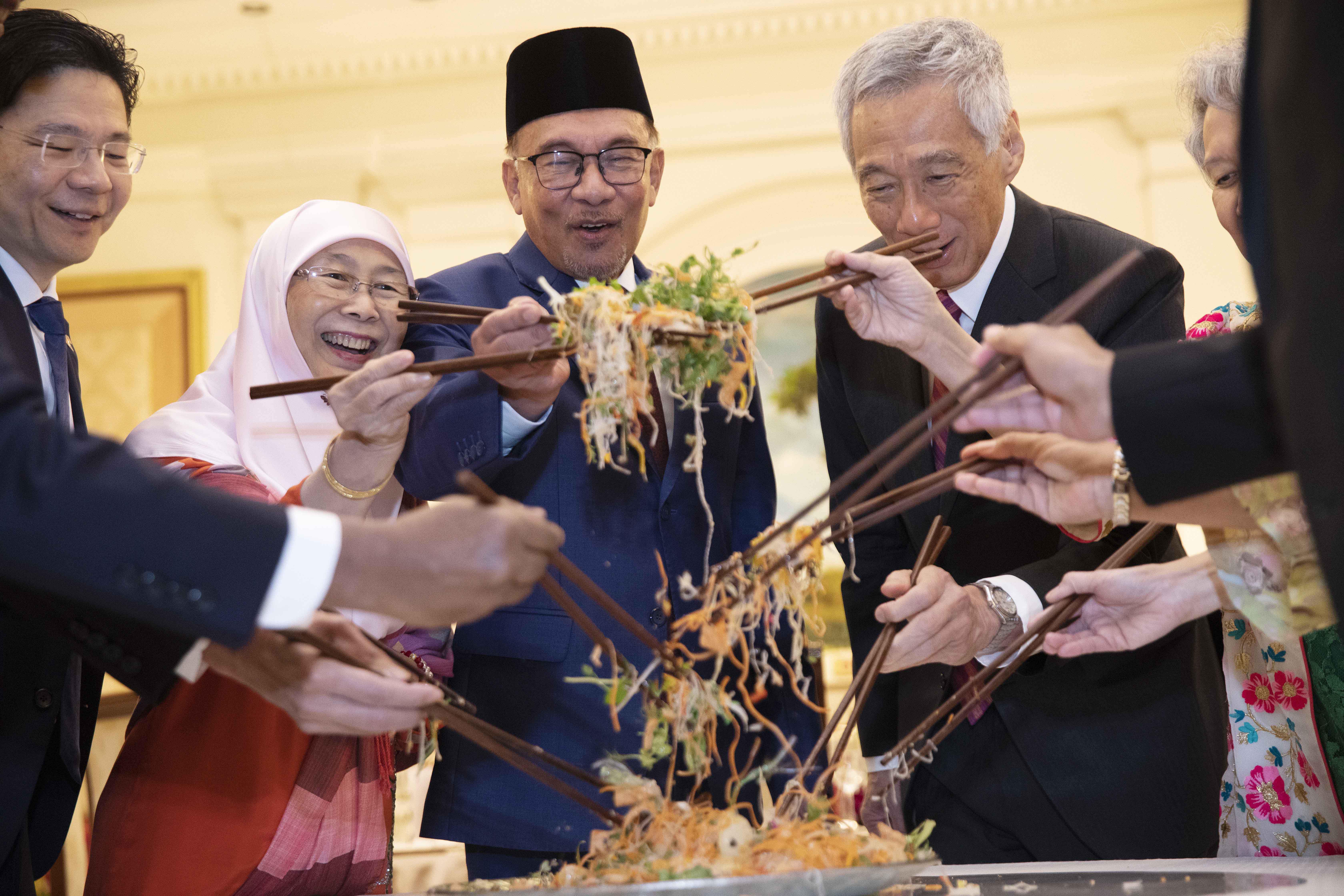 Lawrence Wong tossing 'yee sang' with Wan Azizah Wan Ismail, wife of Malaysian Prime Minister Anwar Ibrahim. Anwar Ibrahim, Singapore Prime Minister Lee Hsien Loong and his wife Ho Ching. They are around a table. The plate of yee sang is in the middie and they are using chopticks to mix the dish.