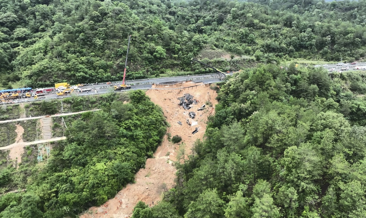 An aerial view of the disaster site. One side of the road has collapsed down a hill. The path left by the collapse is shown by brown earth cutting through the trees
