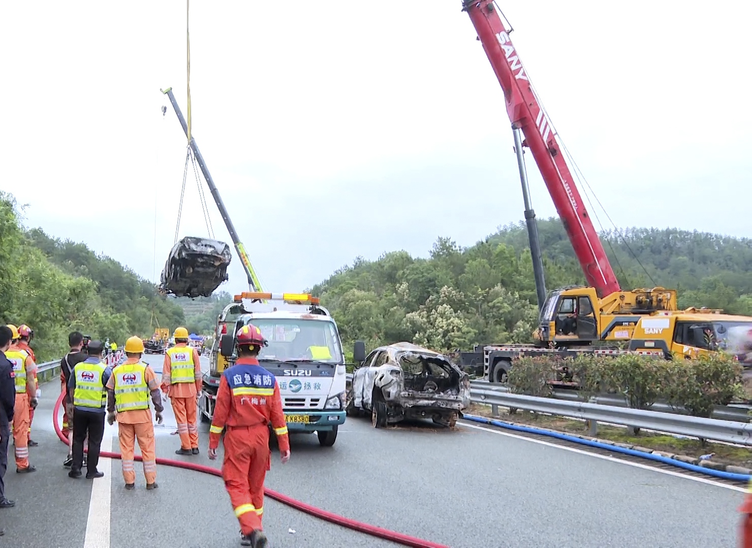 Rescuers at the scene of the collapse. A car is being lifted on a crane above the carriageway. Other recovered cars are already on the road. People in orange unifomrs and hard hats are nearby