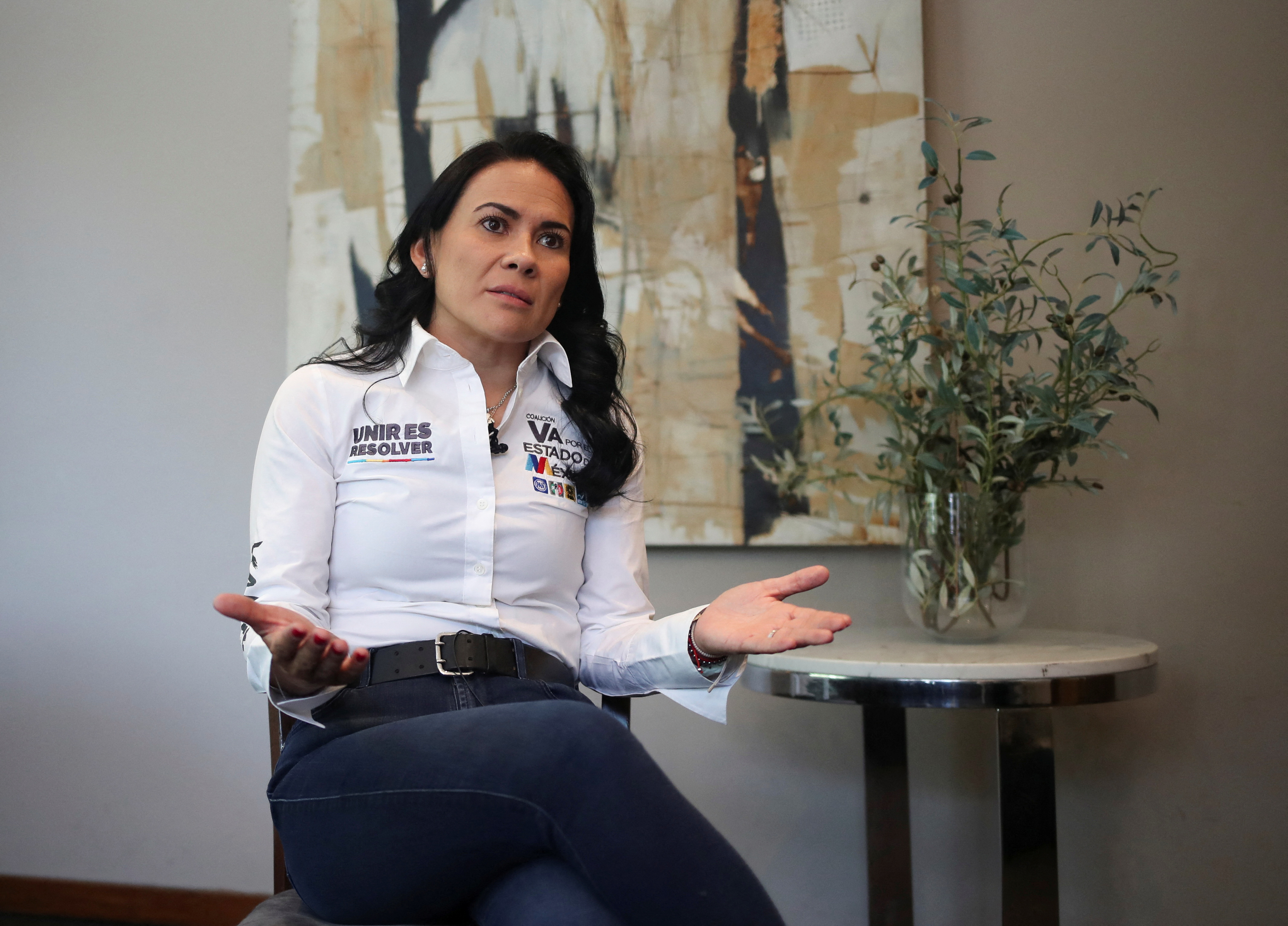 Alejandra del Moral is seated for an interview, her hands outspread in gesture as she speaks. A small table and painting are visible behind her.