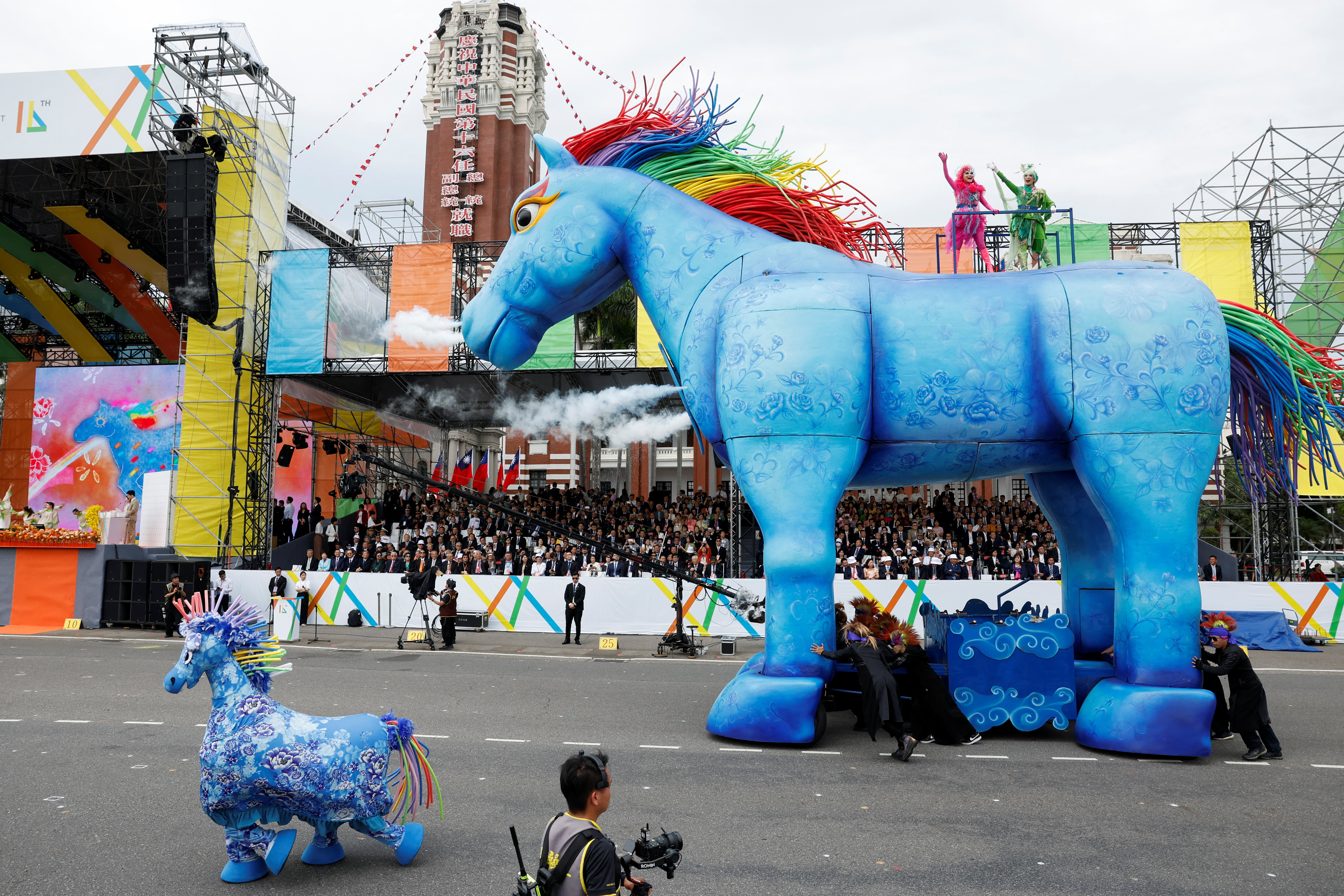 A giant effigy of a blue horse with a rainbow mane participating in the inauguration ceremony. A smaller blue horse is to one side and there are crowds behind