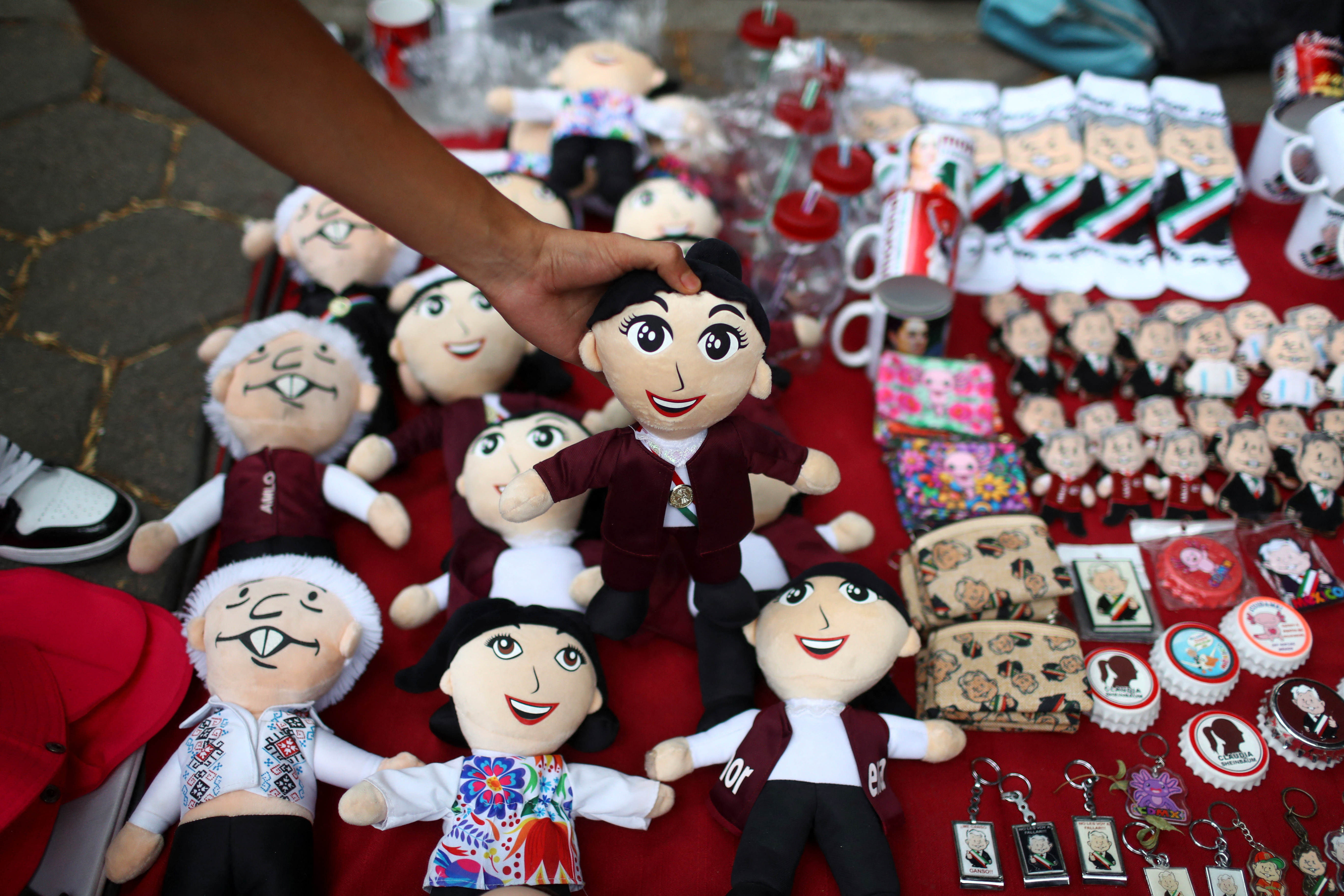 A hand lifts a cloth doll representing Claudia Sheinbaum from a mat covered with election merchandise, including dolls representing Andres Manuel Lopez Obrador.