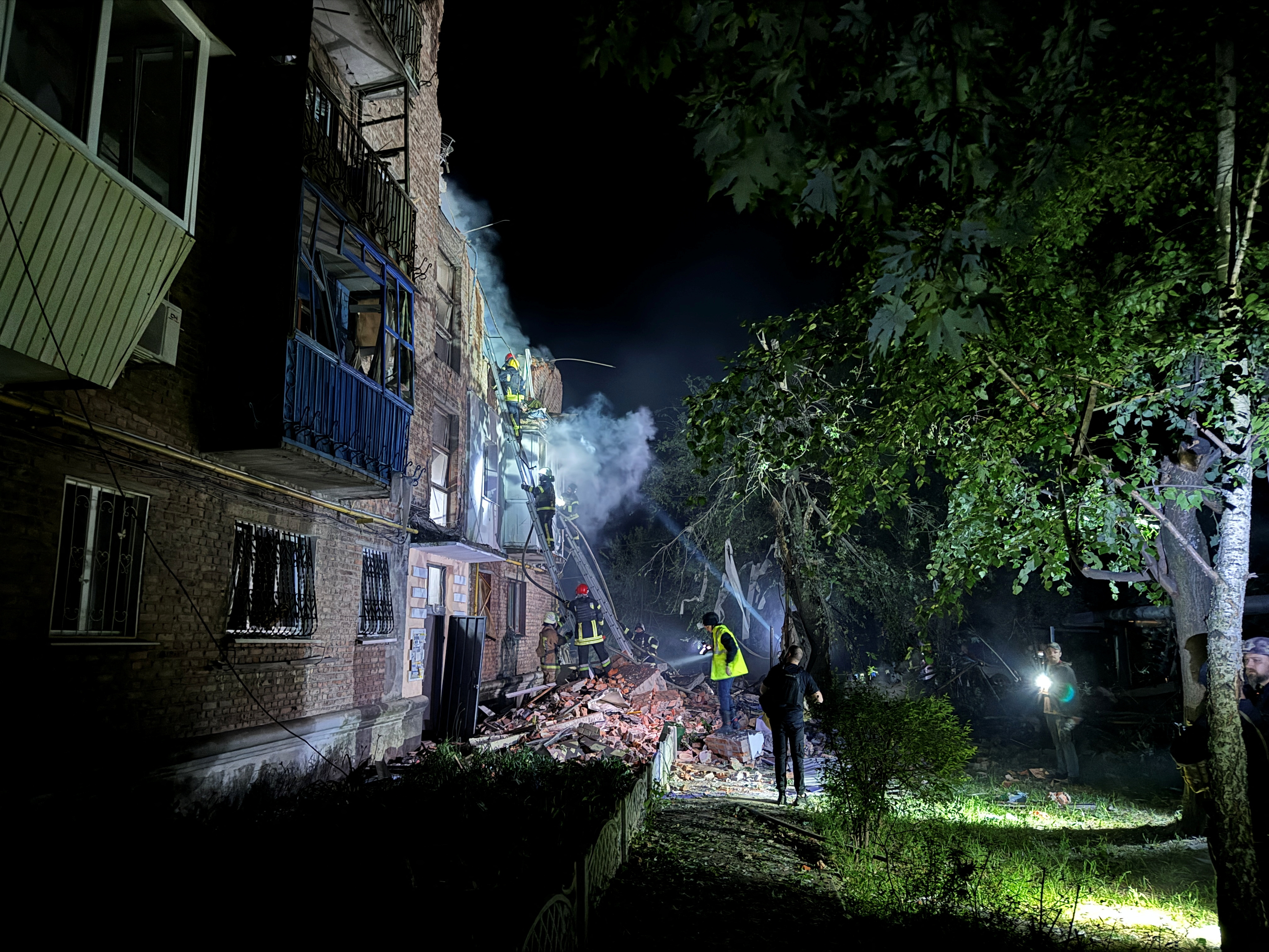 Firefighters at the site of a badly damaged apartment building in Kharkiv after it was hit by a Russian missile. It's night time. The building has been illumintaed. There is a largle pile of rubble, dust and smoke.