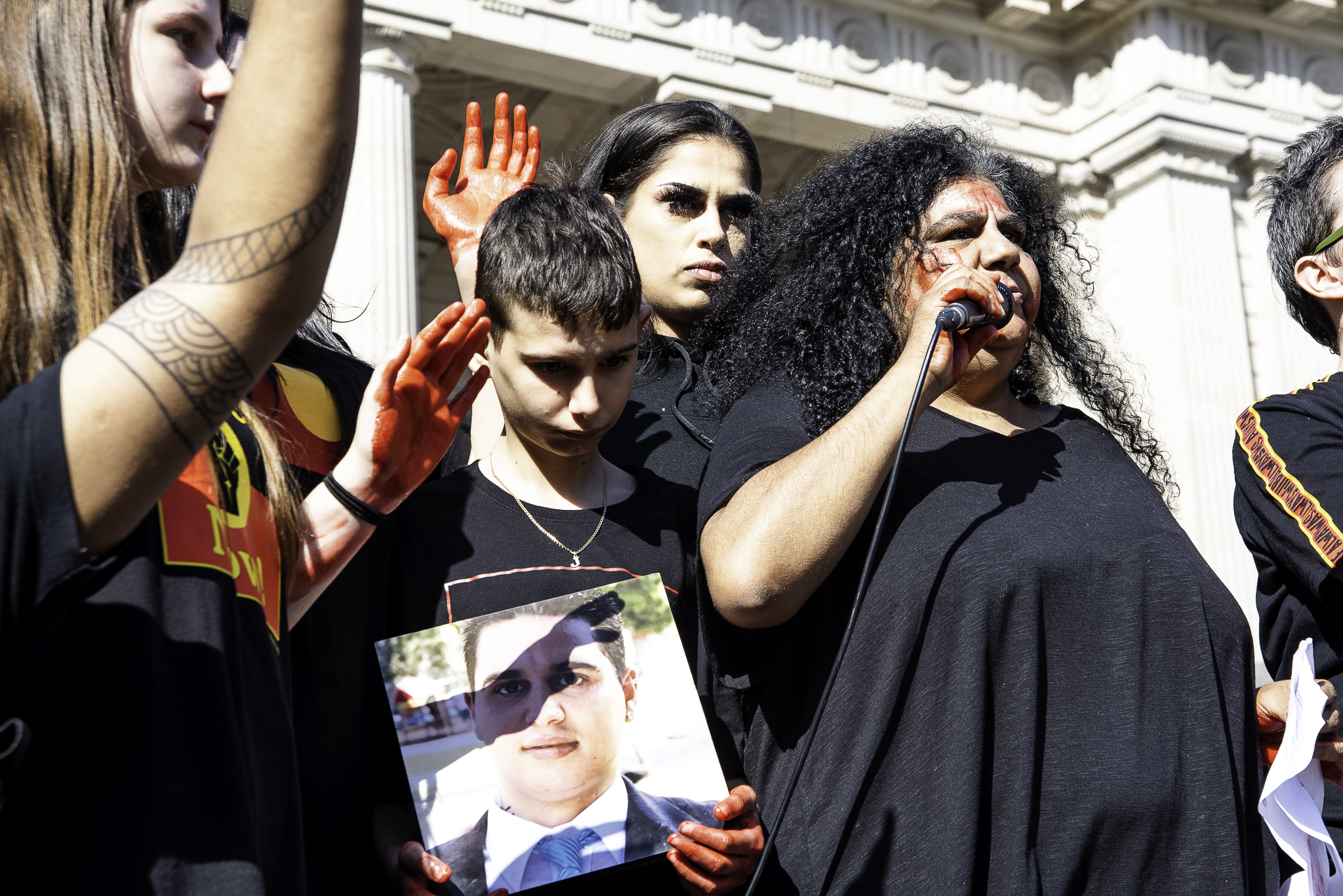 Donnis Kerr. She is at a rally and speaking into a microphone. She has far curly hair and is wearing a black shirt.