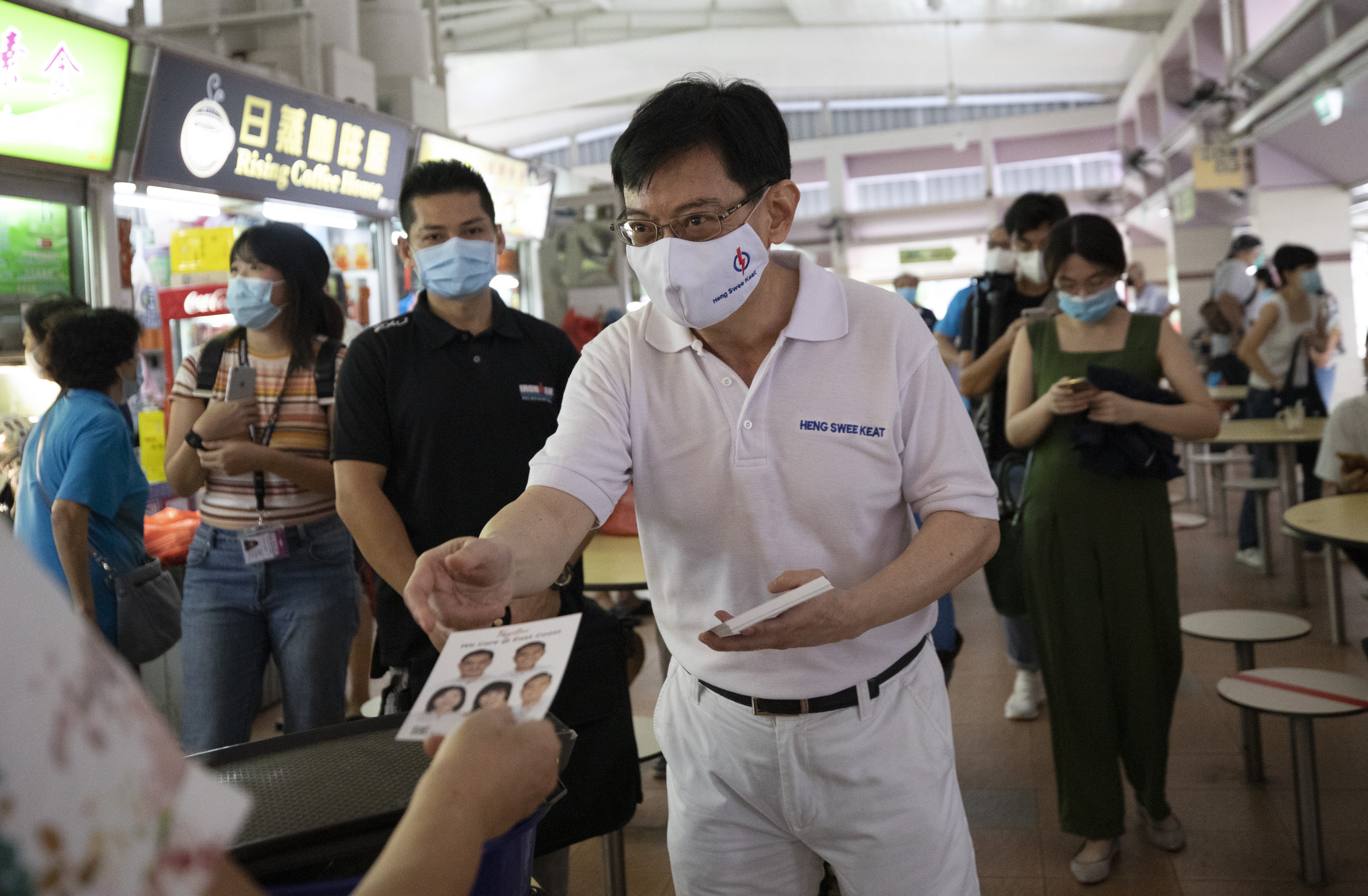 Heng Swee Keat meeting members of the public during the 2020 election campaign. He is wearing white - the colour of the People's Action Party. He is in a hawker centre and there are food stalls nearby. He is handing out leaflets.