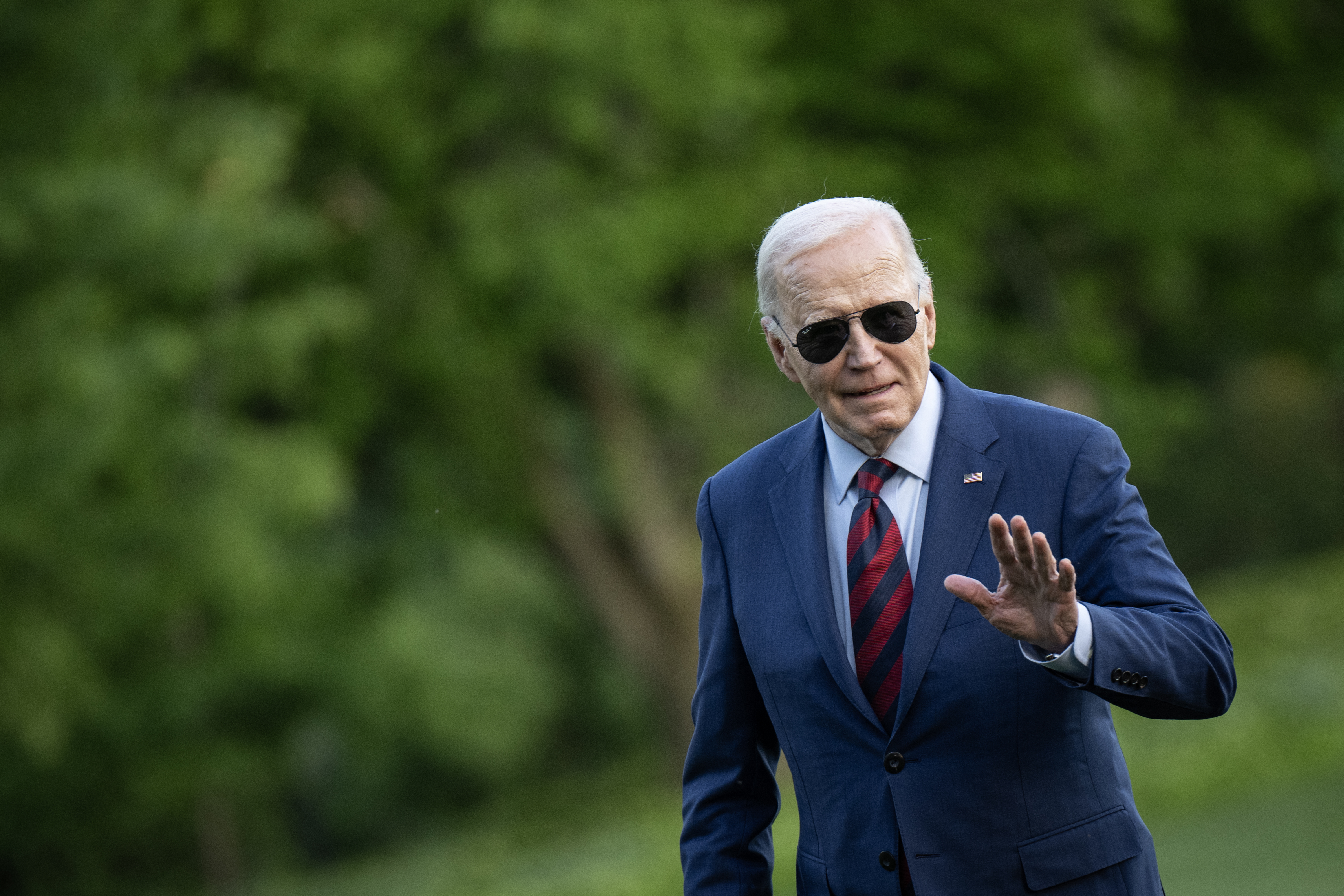 Joe Biden walking across the lawn at the White House. He is wearing a suit and aviator sunglasses. He is waving