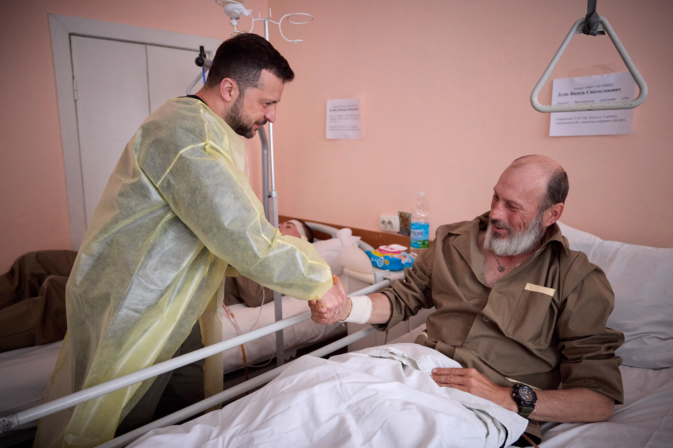 Ukrainian President Volodomyr Zelenskyy greeting an injured soldier in hospital in Kharkiv. Zelenskyy is wearing a yellow medical gown. The soldier is sitting up in bed and shaking hands.