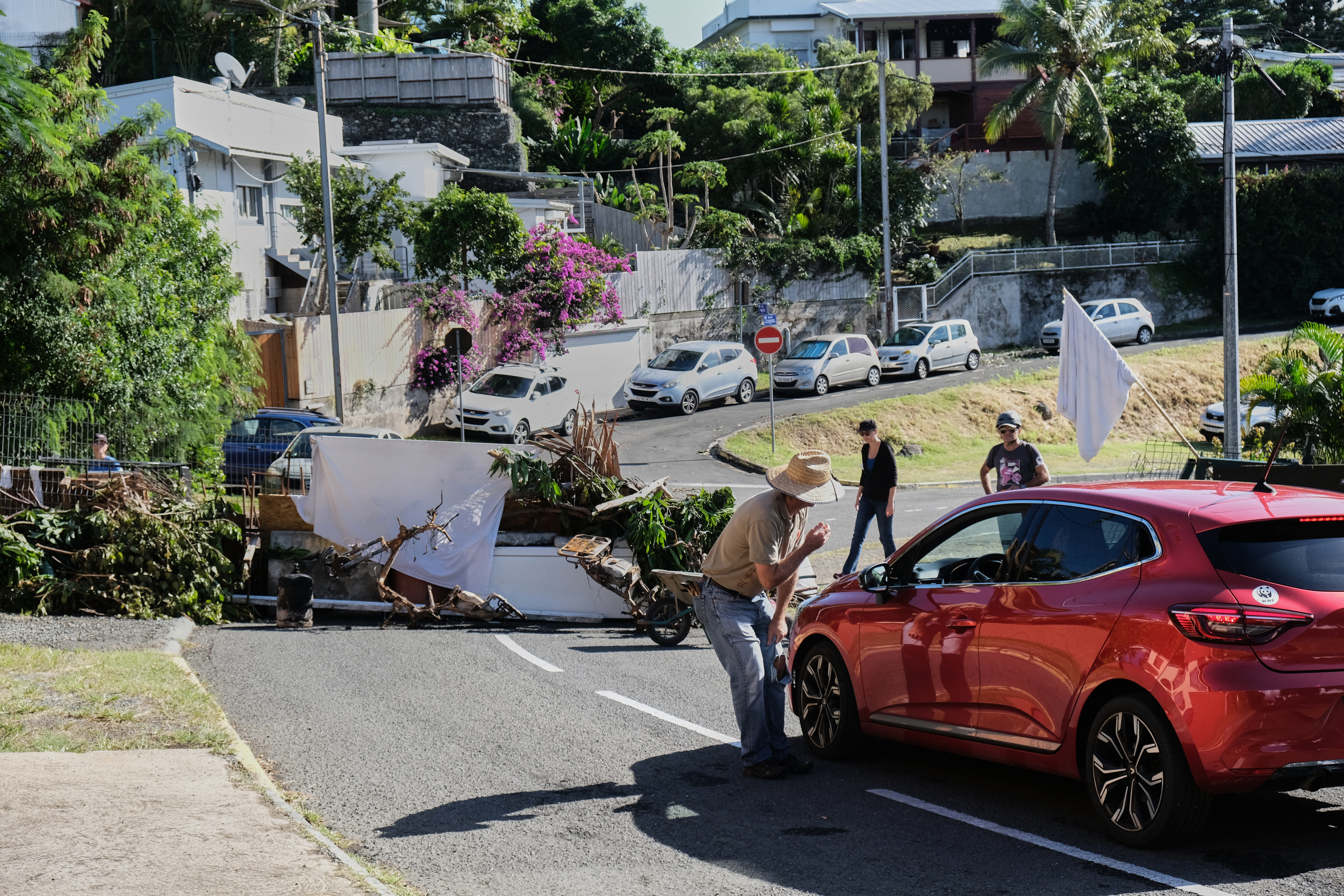 A residents' makeshift barricade in a Noumea district. A man is speaking to the driver of a car. Other people are in the background. There is a white flag. There are houses on the hillside.