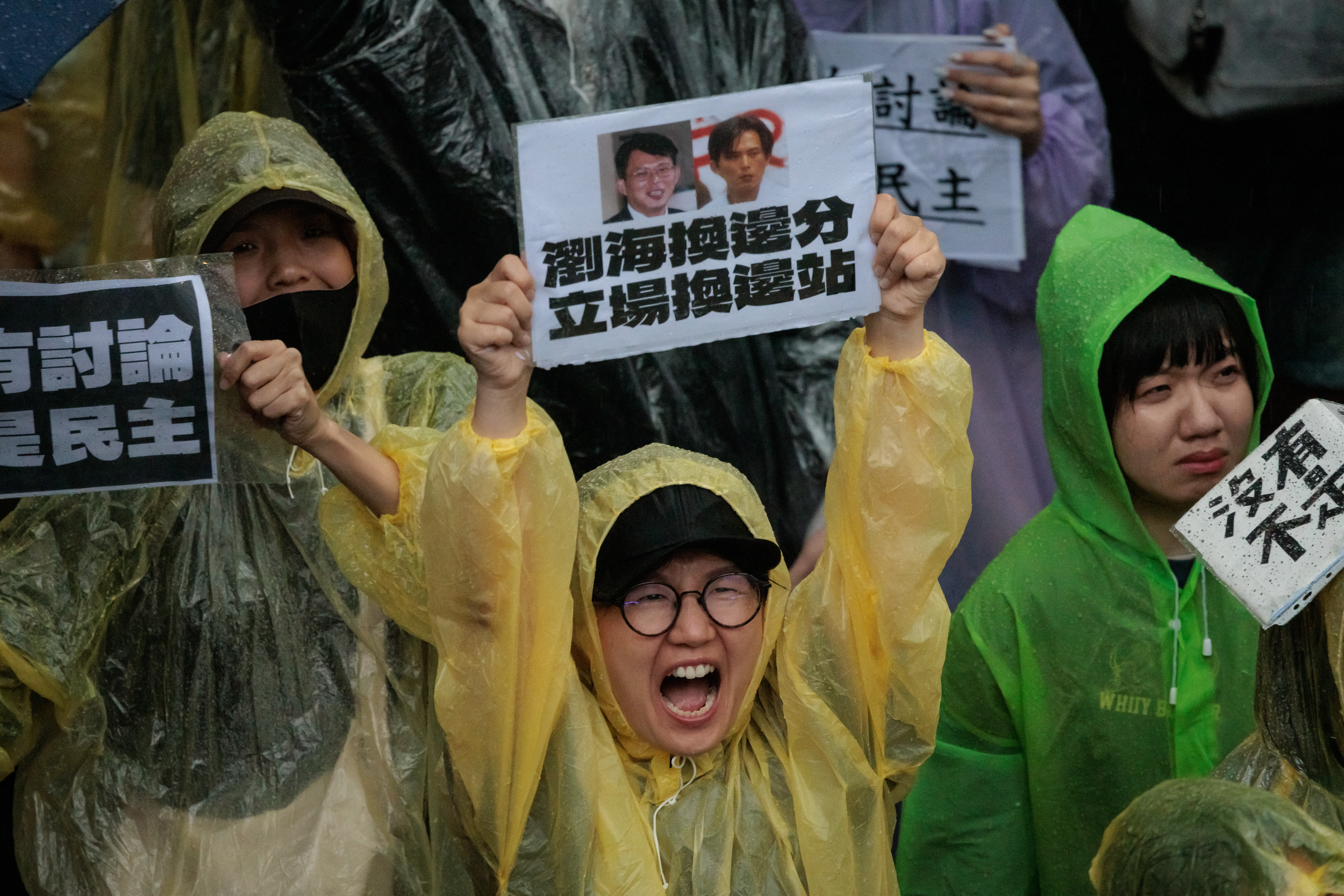 A Taiwanese protester holding up a banner and shouting. Other women are alongside her. They are wearing colourful rain coats.