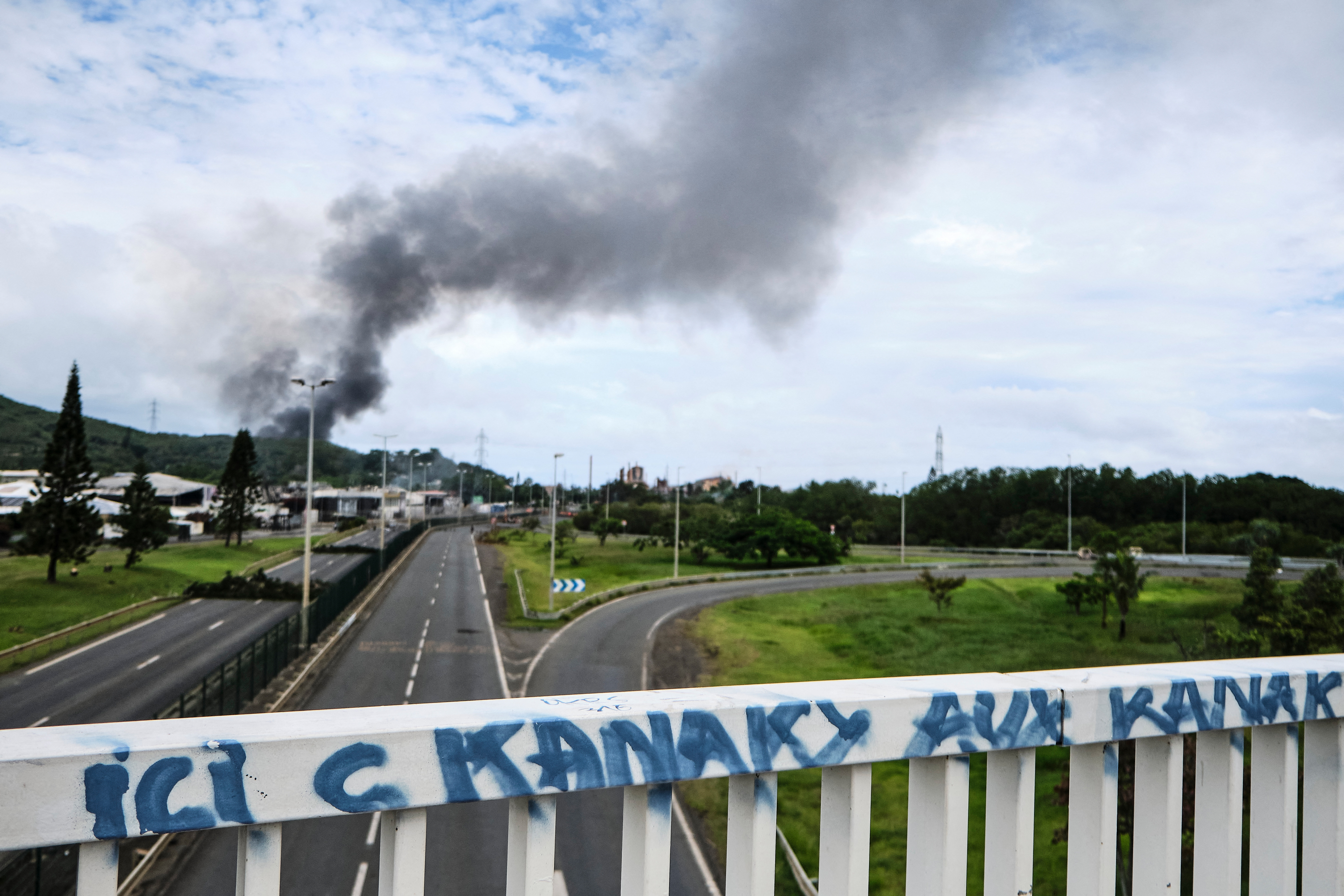 A road bridge with graffiti saying 'Kanaky for the Kanaks' as smoke rises in the distance.