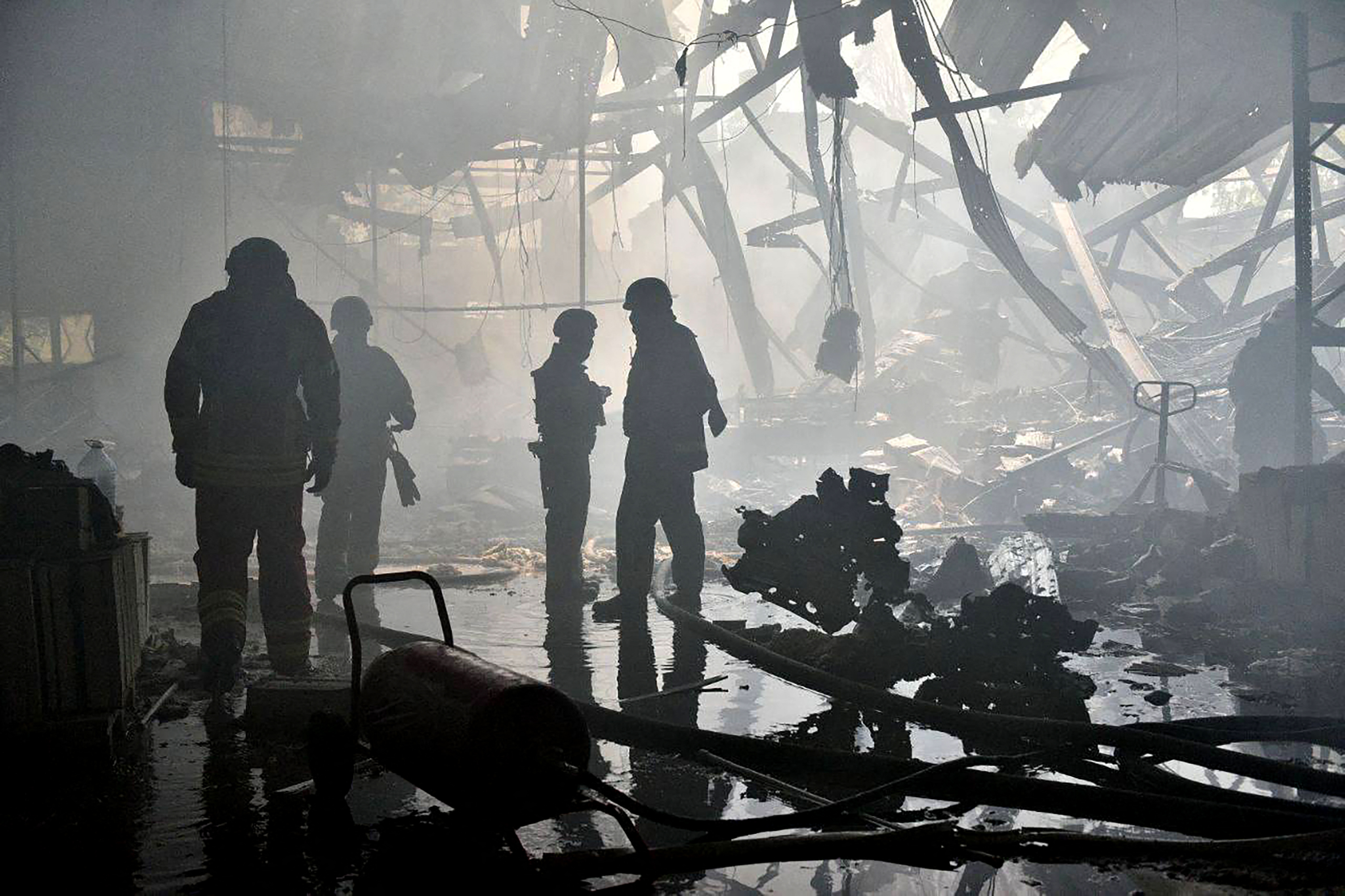 Rescue workers silhouetted through smoke in a destroyed building after a Russian missile attack on Kharkiv. There ia.a large hole in the ceiling and debsri on the floor and hanging down.