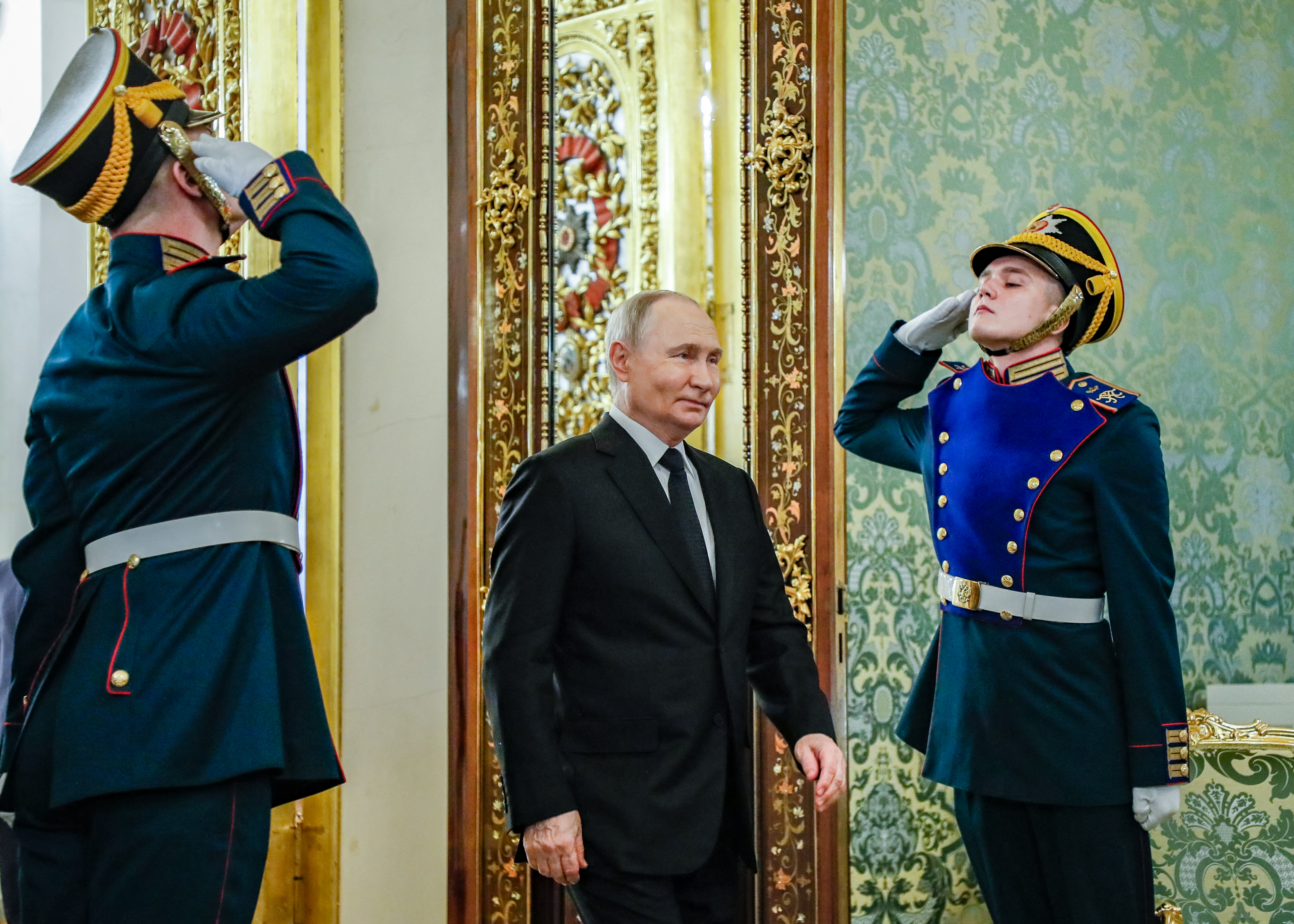 Putin being saluted by two ceremonial guards in the Kremlin. The room looks palatial with tall, gilded doors.