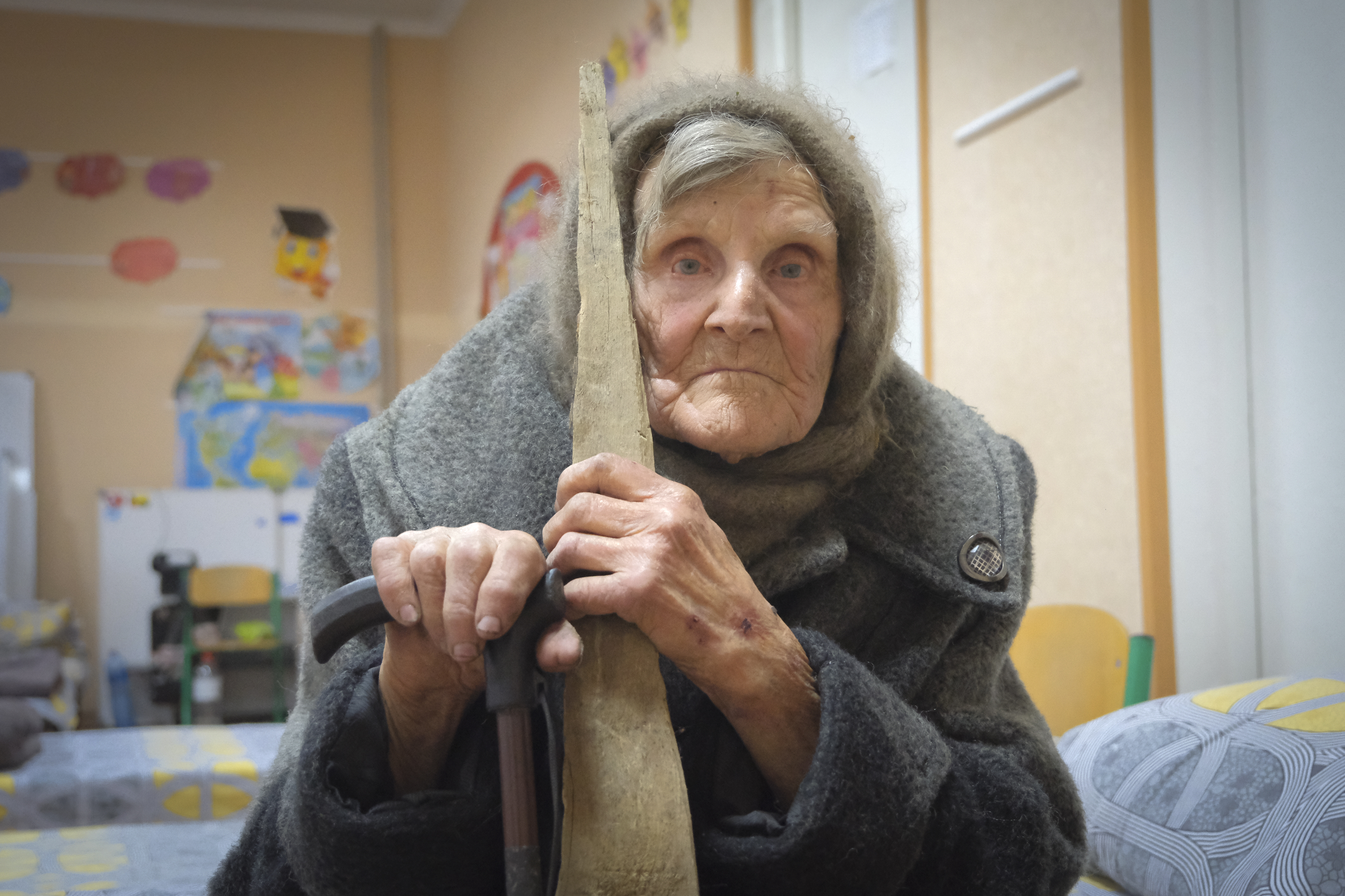 Lidya Stepanovna. She is sitting on a bed with a shawl around her head and leaning on a cane.