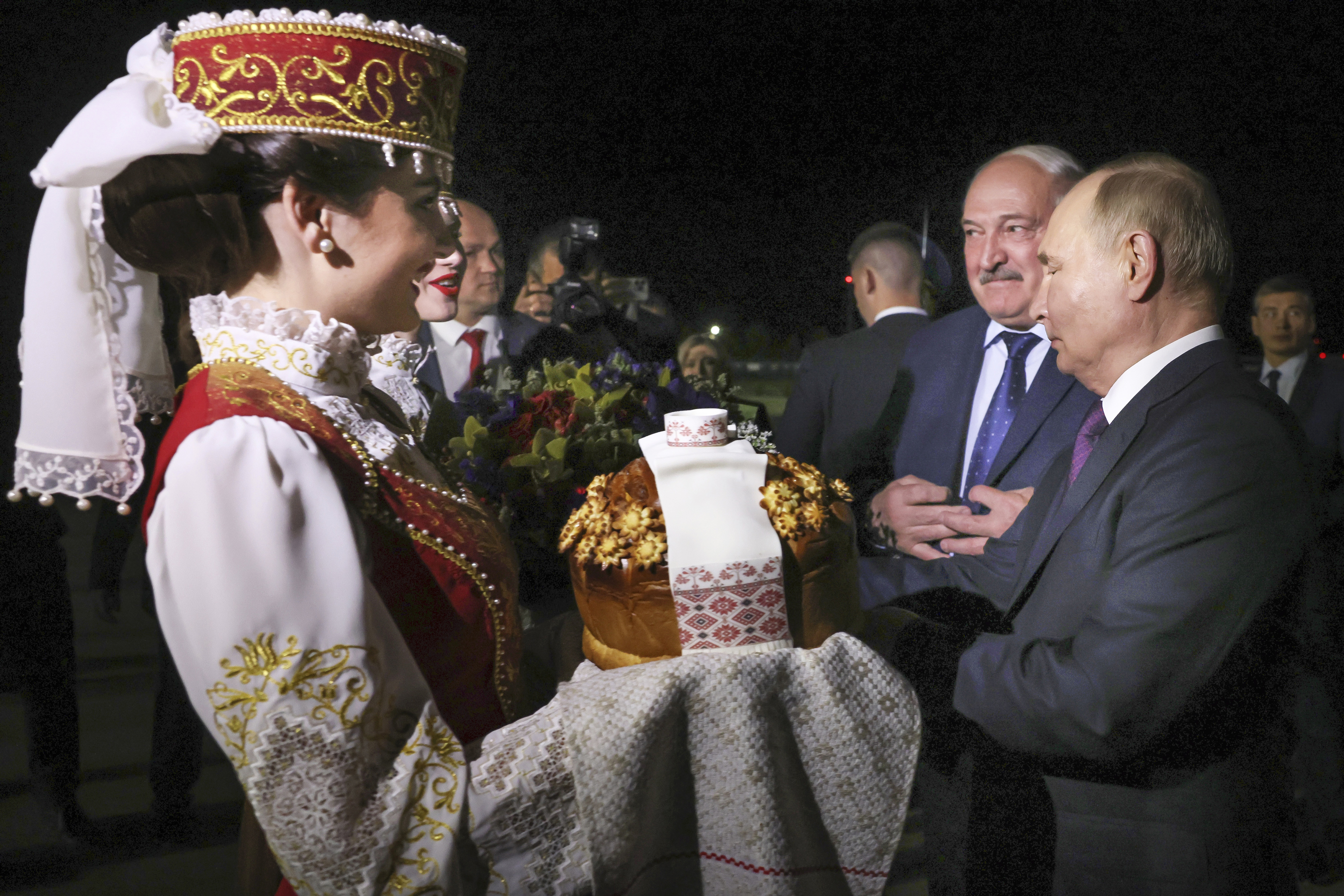 Alexander Lukashenko welcoming Vladimir Putin to Belarus. There s a woman in traditional dress on the left holding a gift. A woman behind her has a bouquet of flowers. Lukashenko and Putin are on the right.