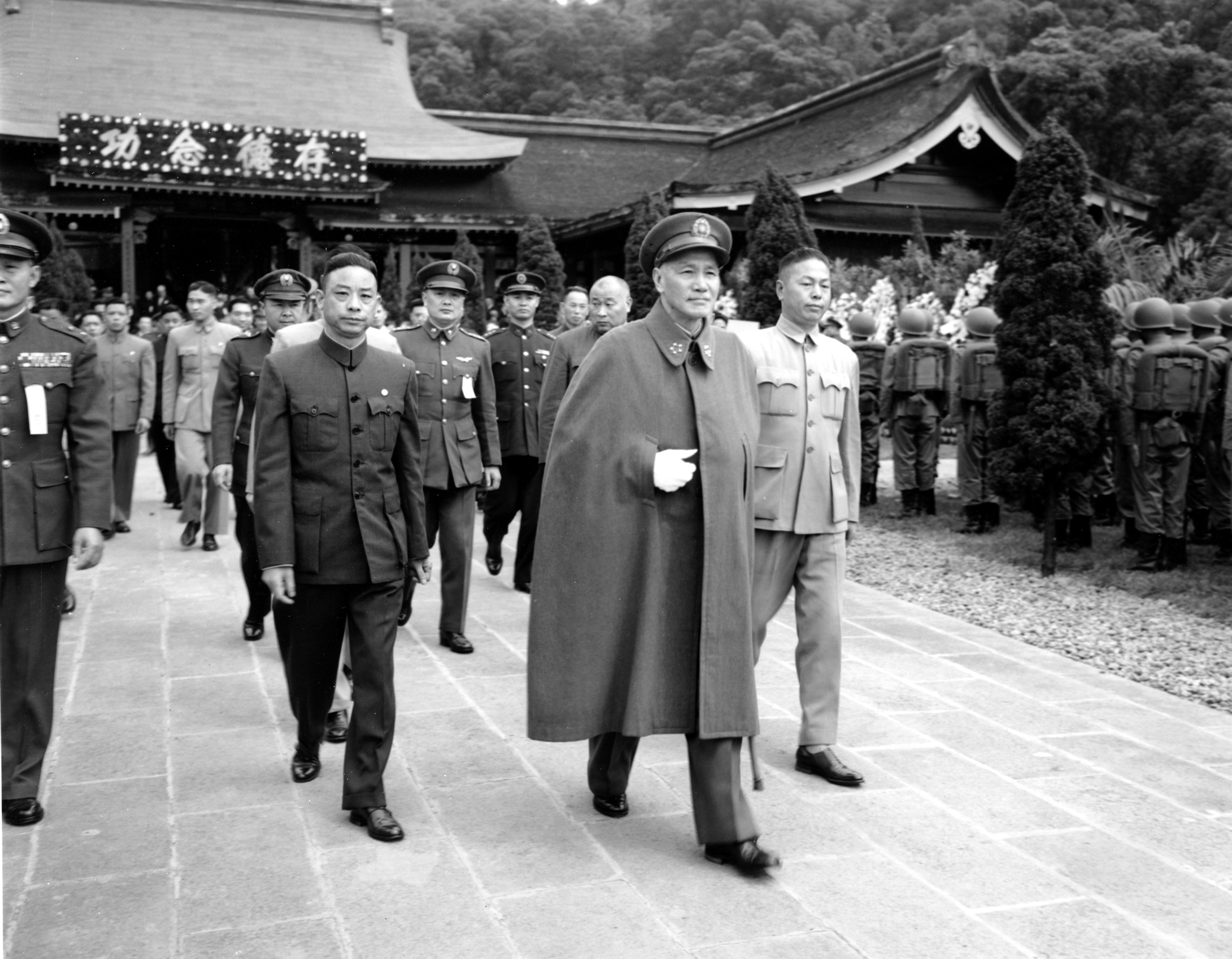 Chiang Kai-shek pictured in 1955. He is wearing a military uniform with a long cape. Others in uniforms are walking behind him. They are leaving a temple.