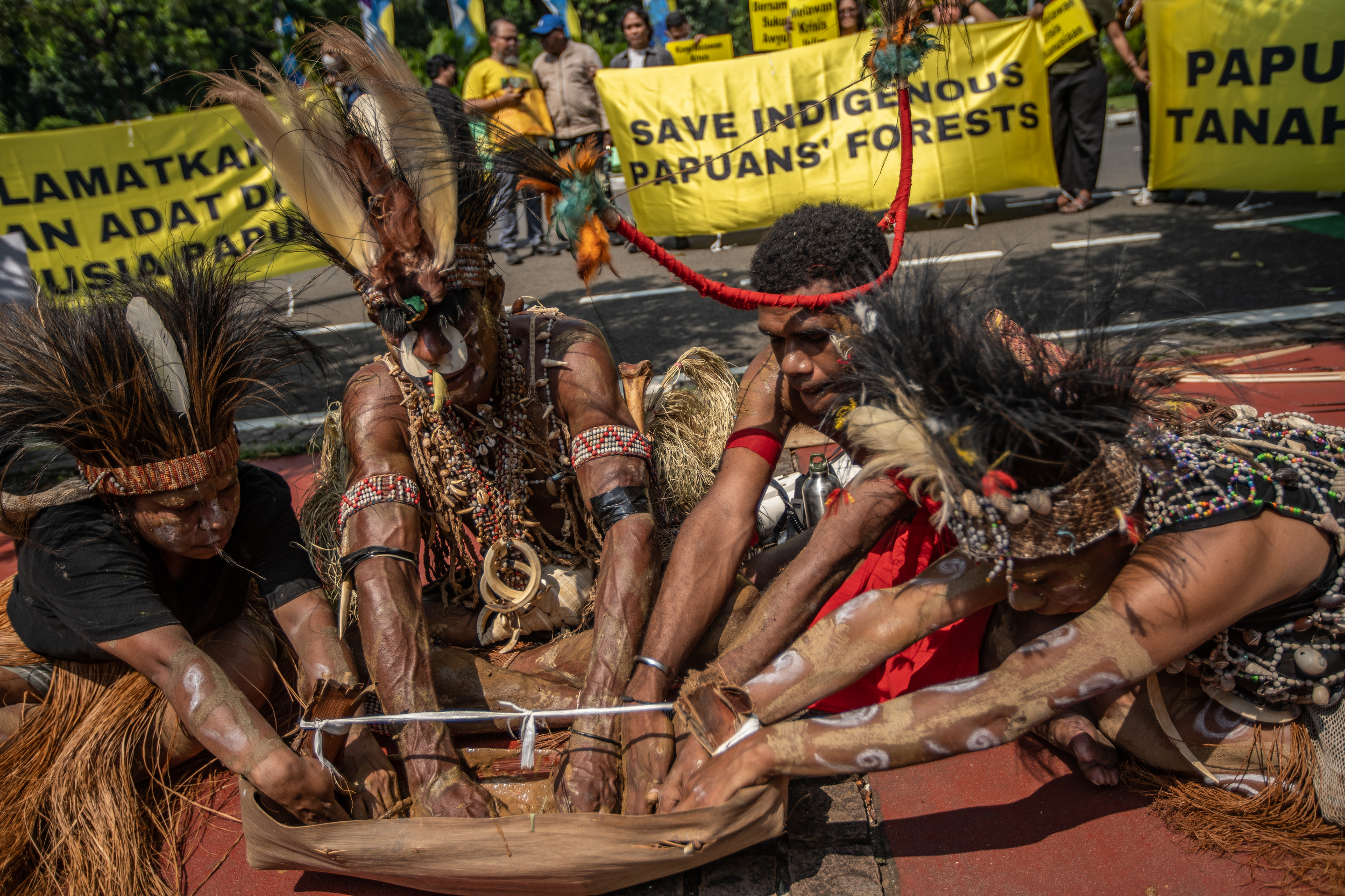 Indigenous people from West Papua sitting around a bark container with the soil of their homeland. The container is in the middle and they are seated around it. Their hands are outstretched.
