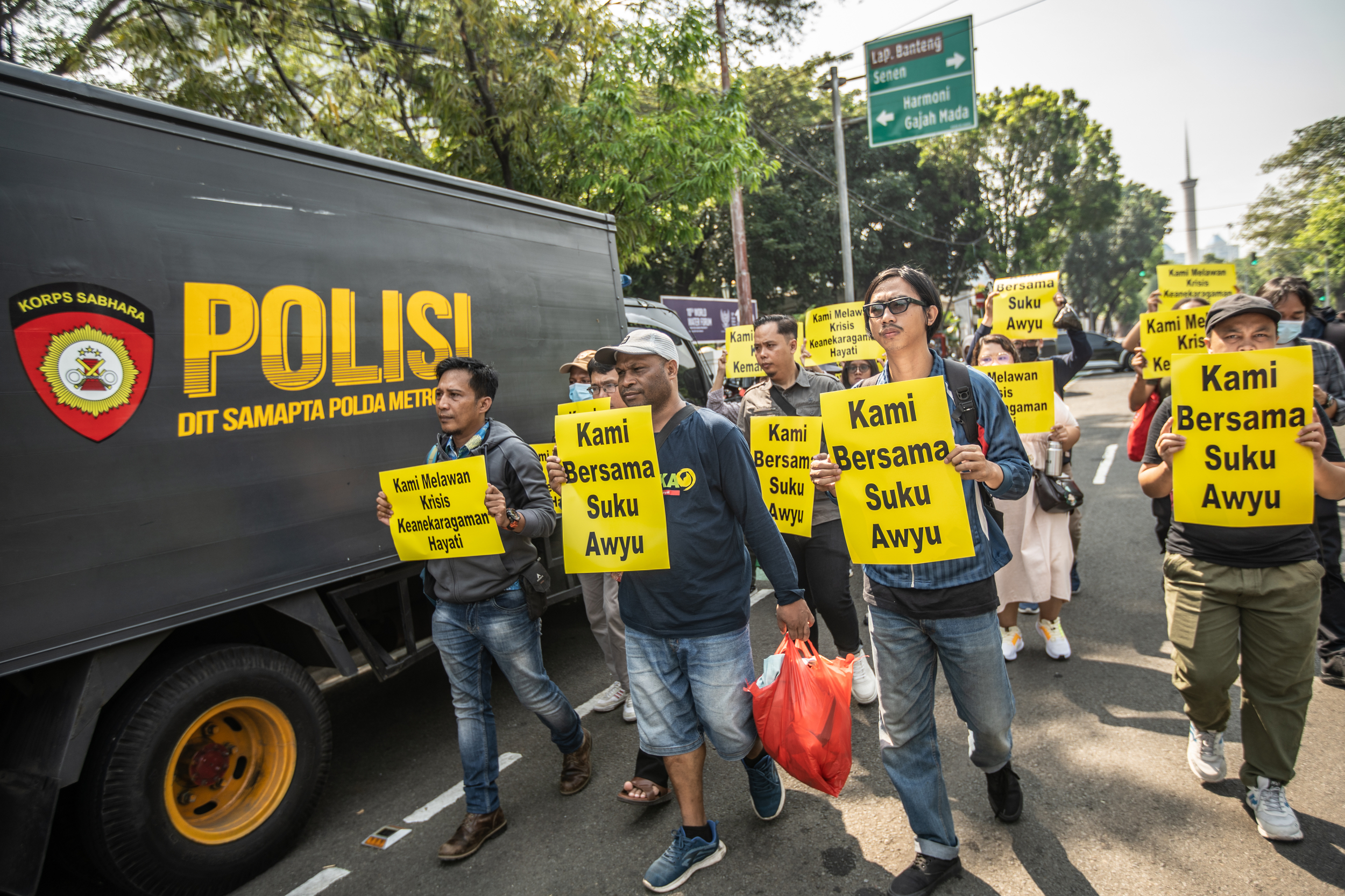 Protesters carrying yellow placards reading 'Kami Bersama Suku Awyu' {We're with the Awyu people'] They are walking next to a police truck