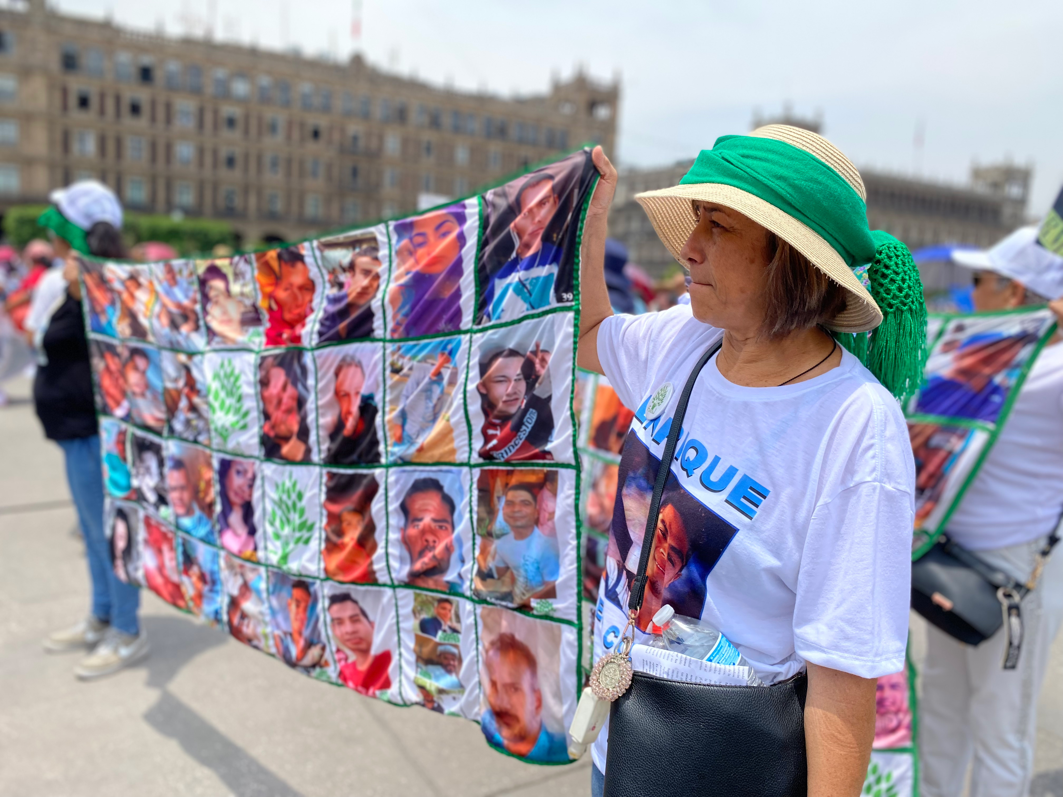A woman in a straw hat holds up one side of a large banner that shows the faces of missing people in Mexico. Her T-shirt is likewise printed with the face of a man, Enrique, who has gone missing.