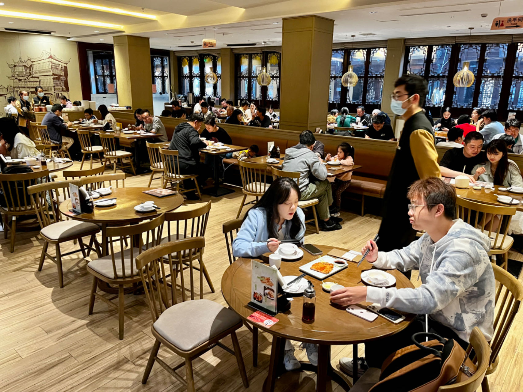 The dining room of the Nanxiang restaurant on a Saturday night. Theree couples and families enjoying their dinner. A waiter is standing nearby. One table is empty.