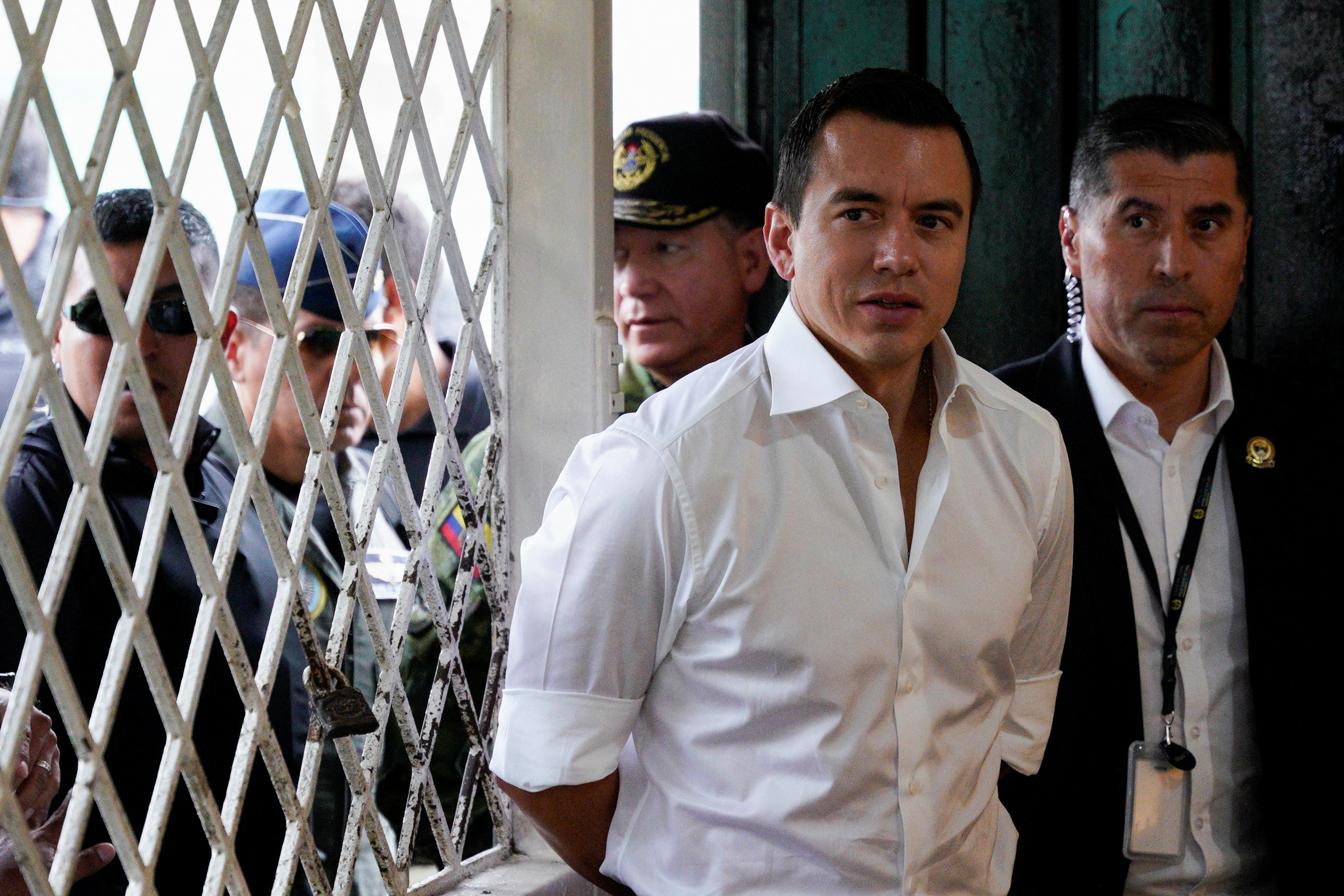 Daniel Noboa — dressed in a crisp, white collared shirt — stands near a white security gate, surrounded by officers.