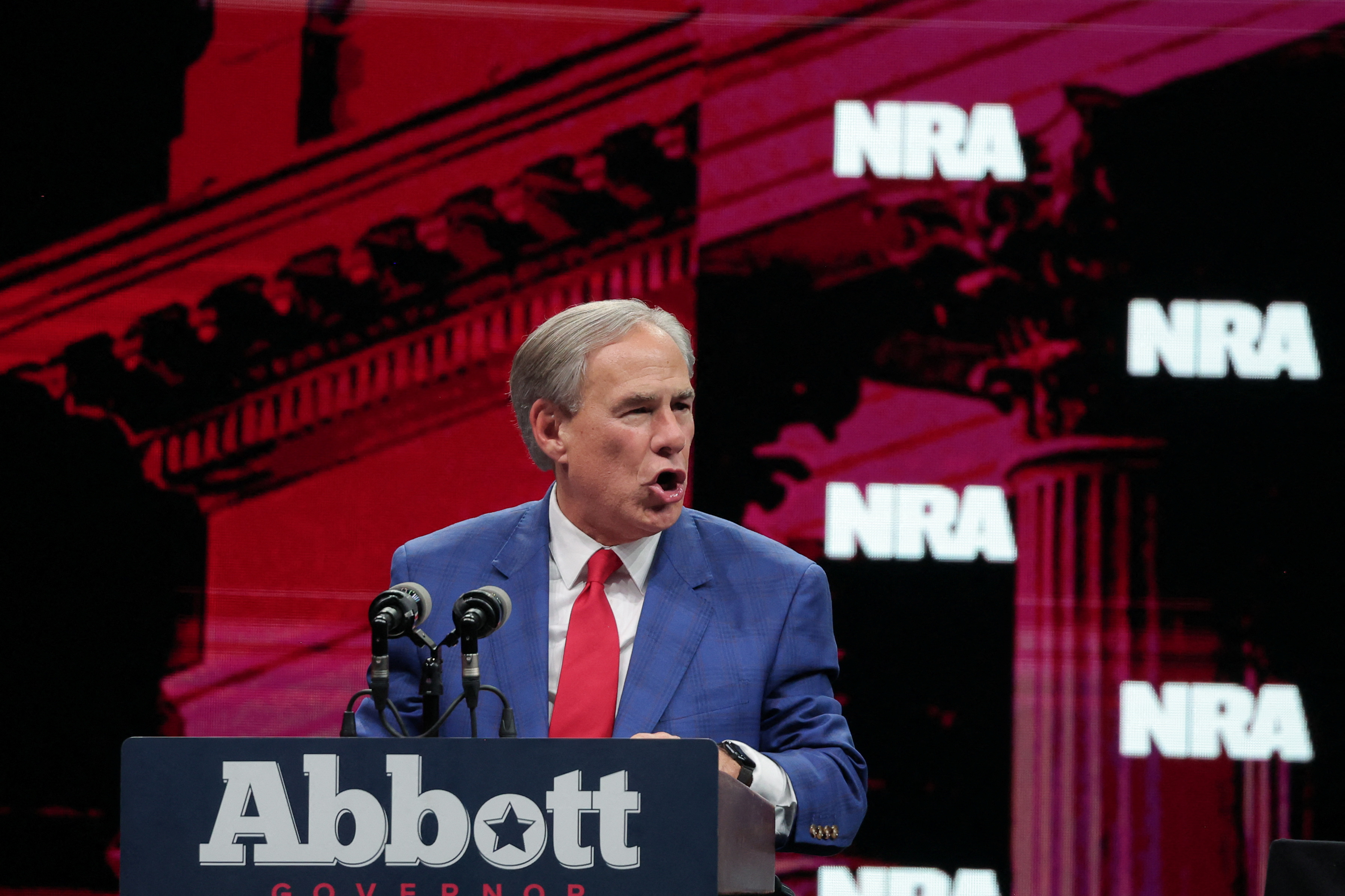 Greg Abbott speaks from behind a podium that bears his name. Behind him is a screen with the letters NRA.