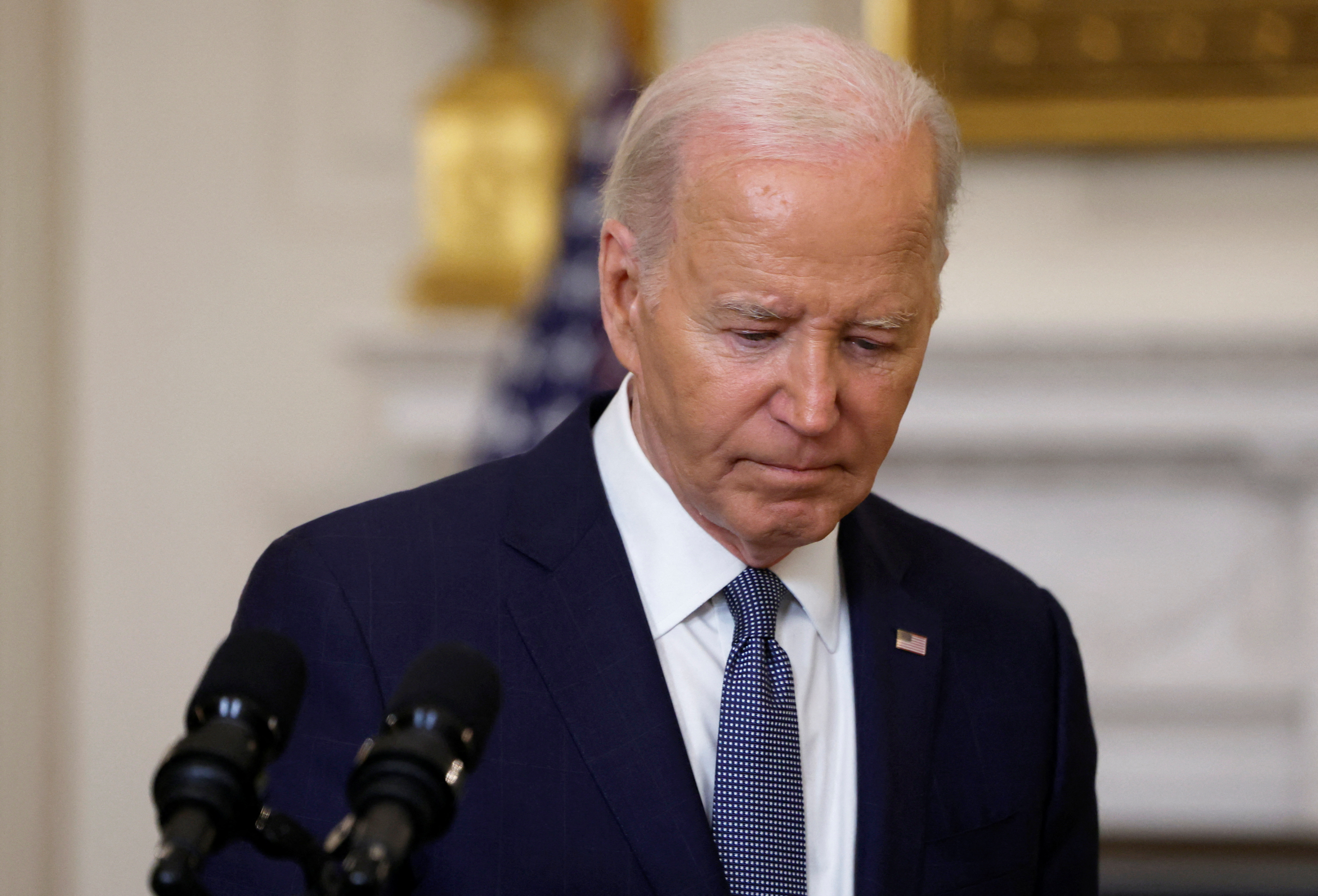 President Joe Biden, looking downward, wears a dark suit and blue tie as he stands in the White House.