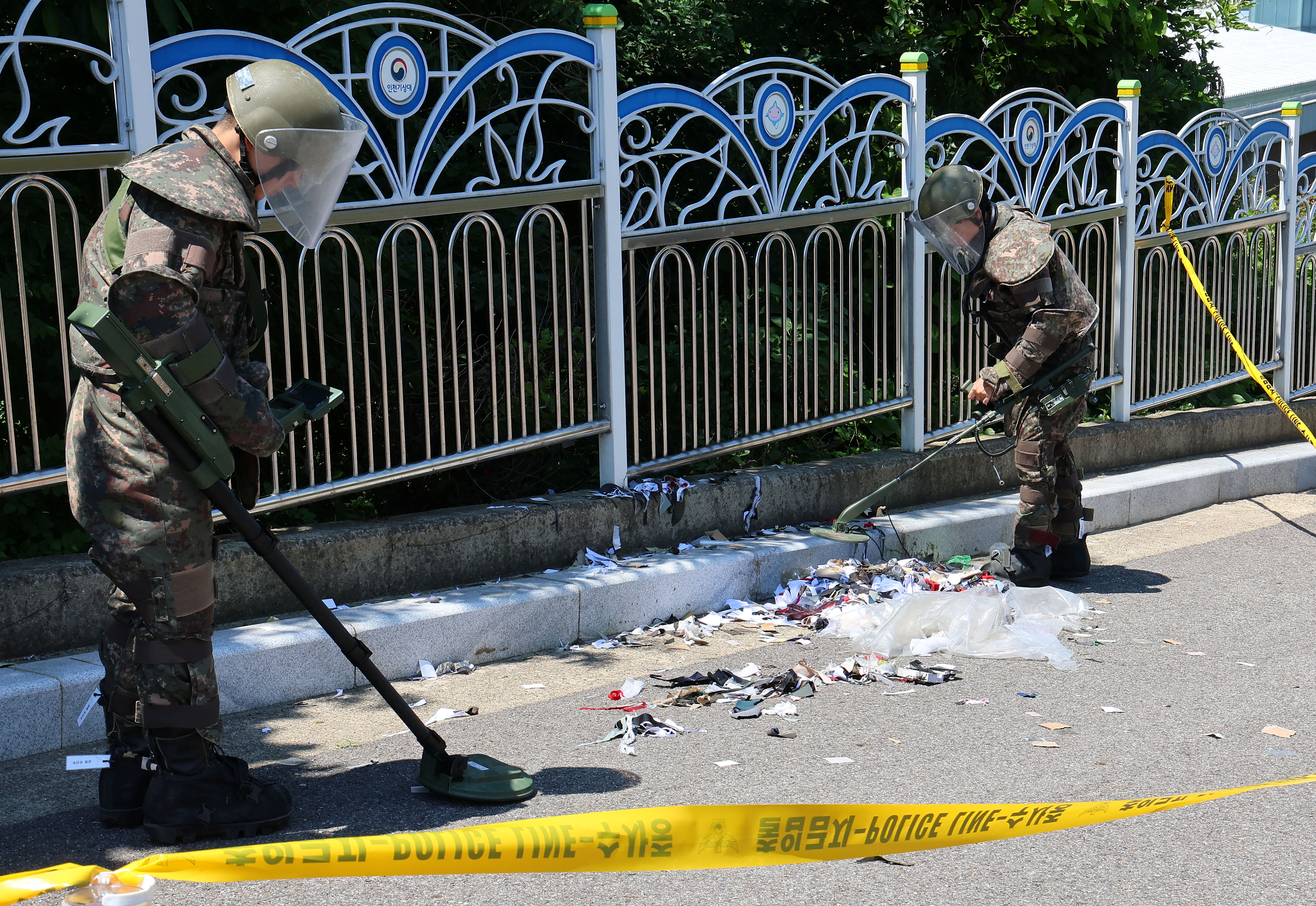 South Korean soldiers looking at debris from a North Korean balloon. They have visors and protective suits. The area is cordoned off behind yellow tape.