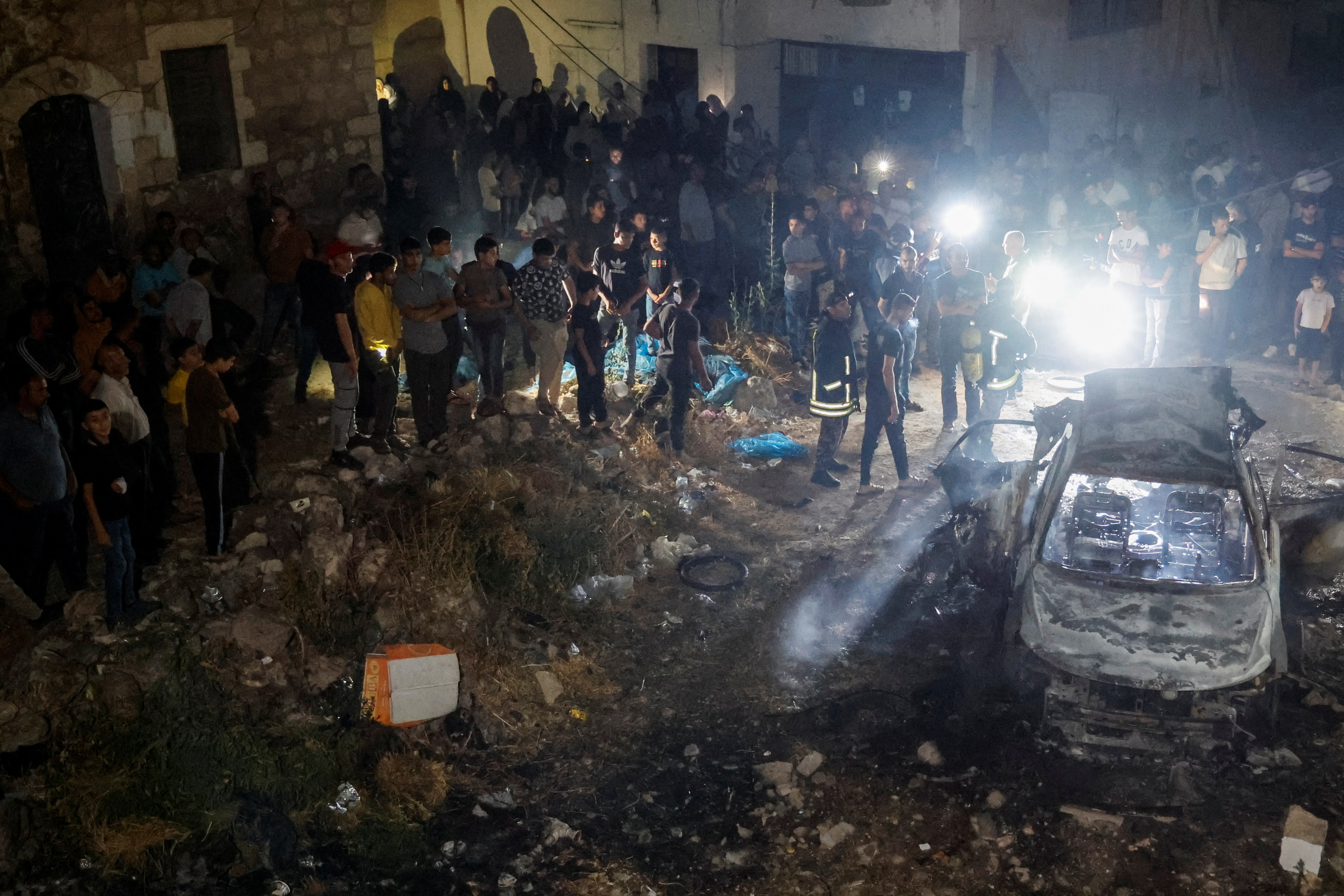 People and rescuers gather around a burnt out car at night