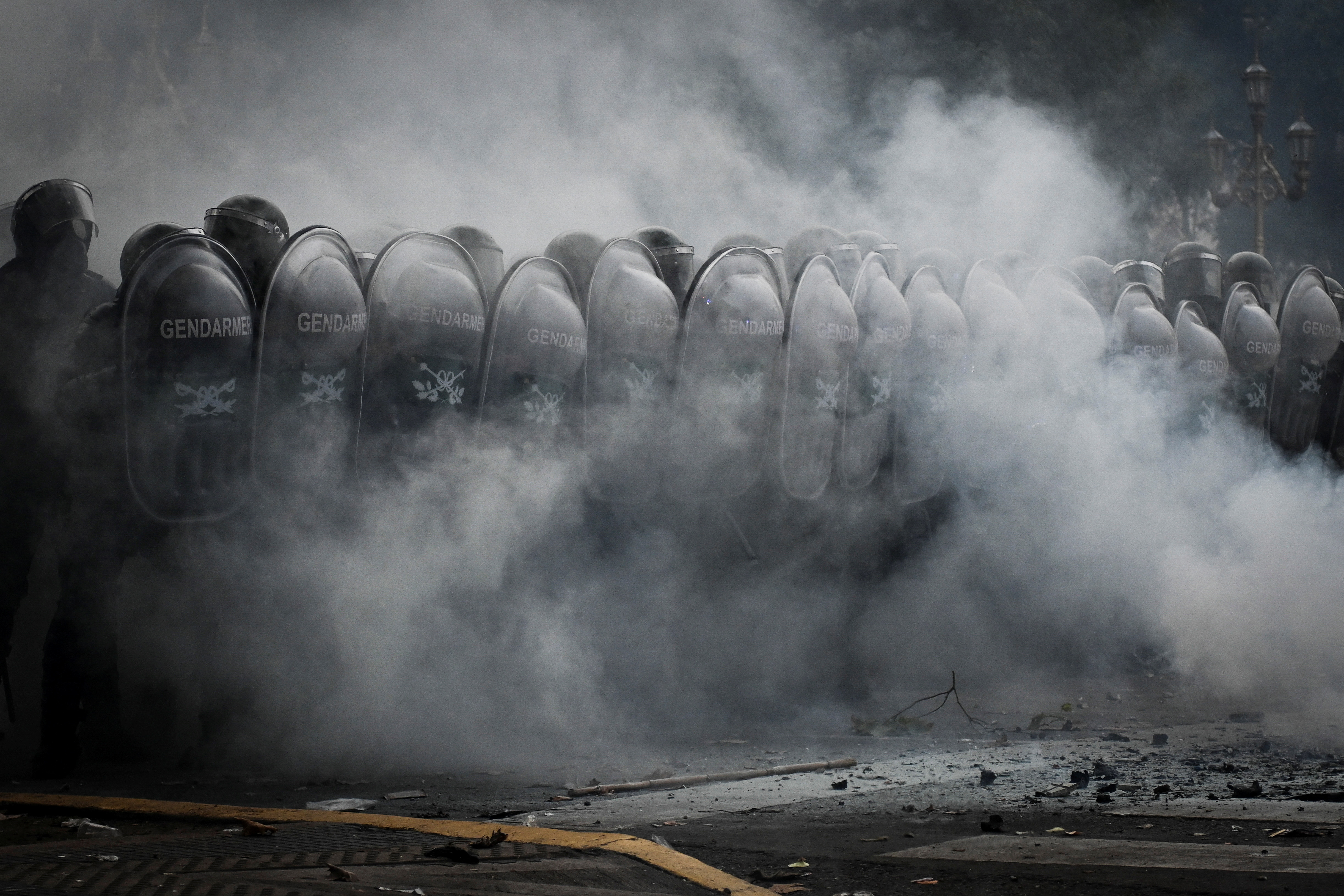 A row of police behind clear riot shields, obscured by a cloud of what is likely tear gas.