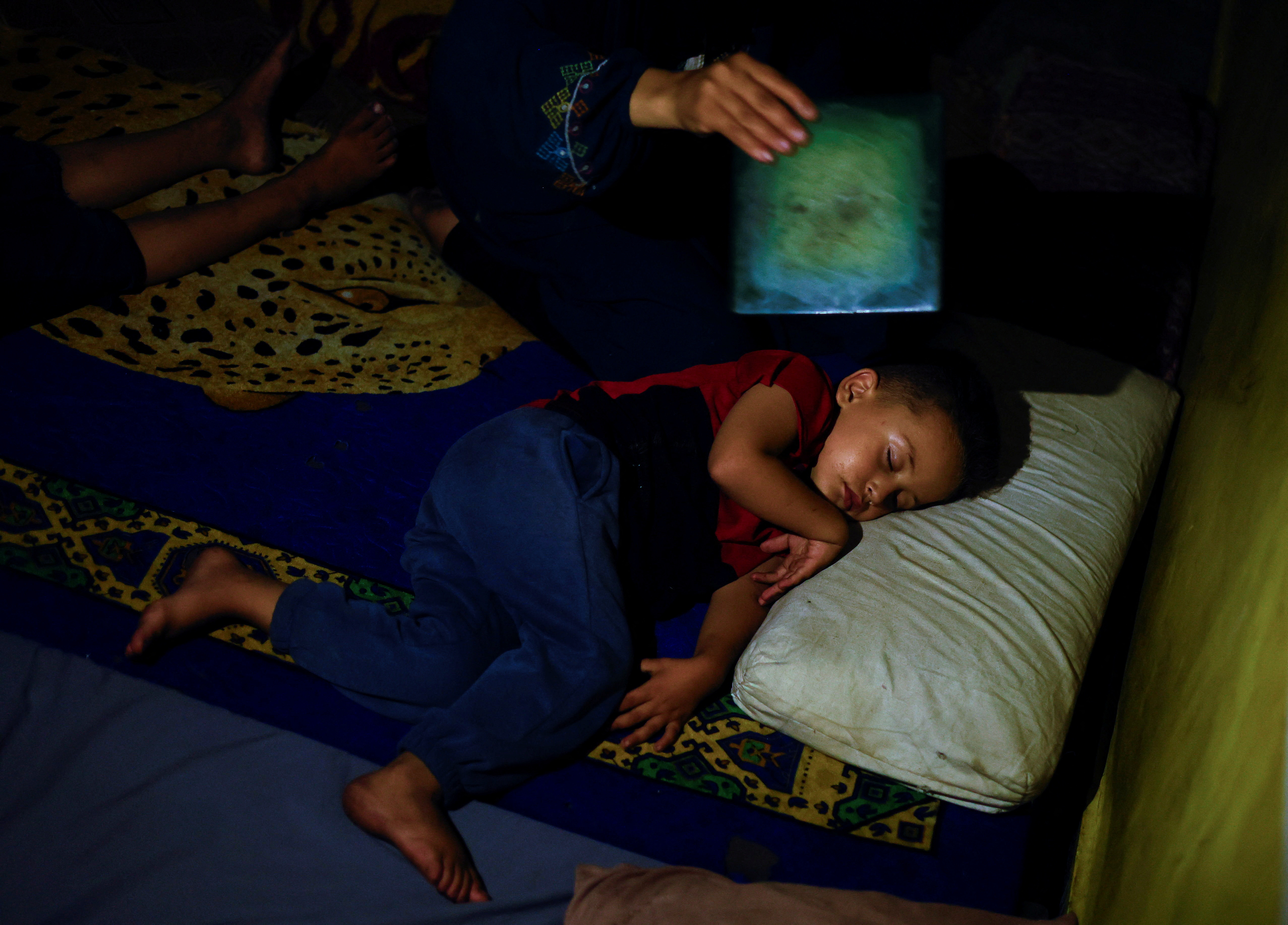 A displaced Palestinian woman, Amal Nsair, fans her son as he sleeps in a school where they shelter, during hot weather and power cut