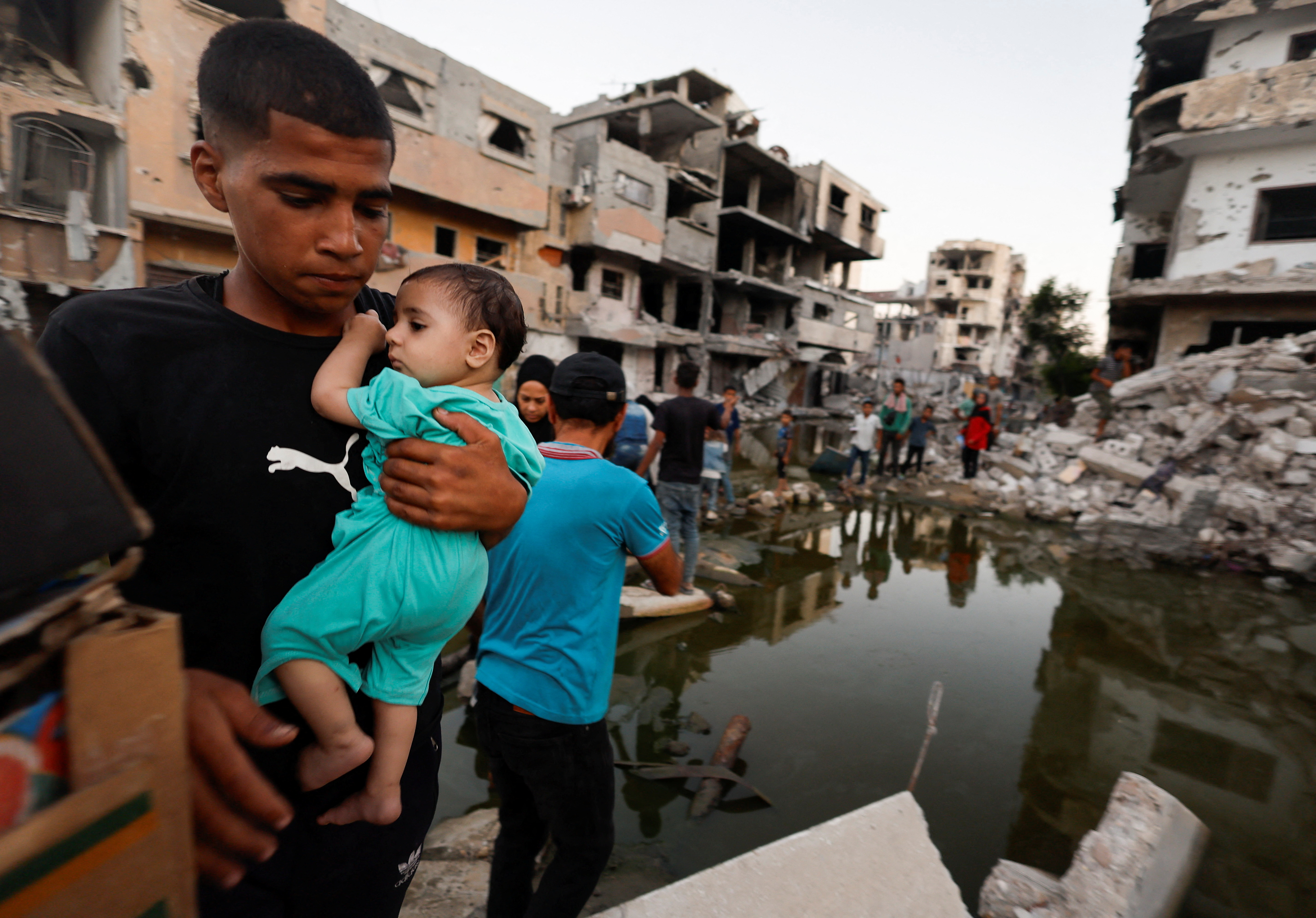 A boy holds a baby as people walk through sewage in a destroyed city