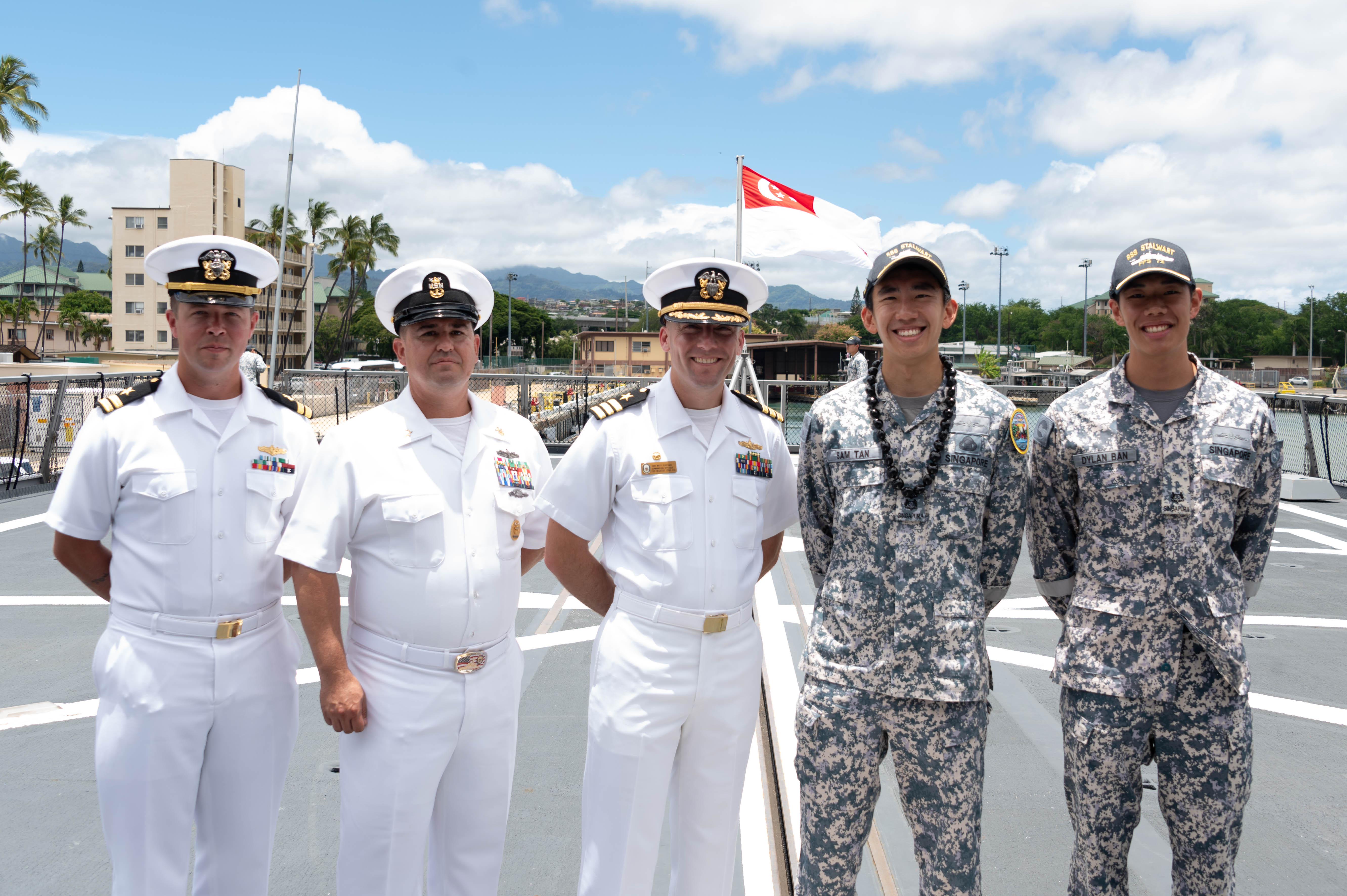 Two Singaporean sailors from the frigate RSS Stalwart standing alongside three US Navy sailors on the ship's deck after they arrived in Hawaii. Singapore's naval ensign is flying behind them. The Singapore sailors are in combat uniform and wearing baseball caps with the ship's name. The US sailors are in white uniforms.