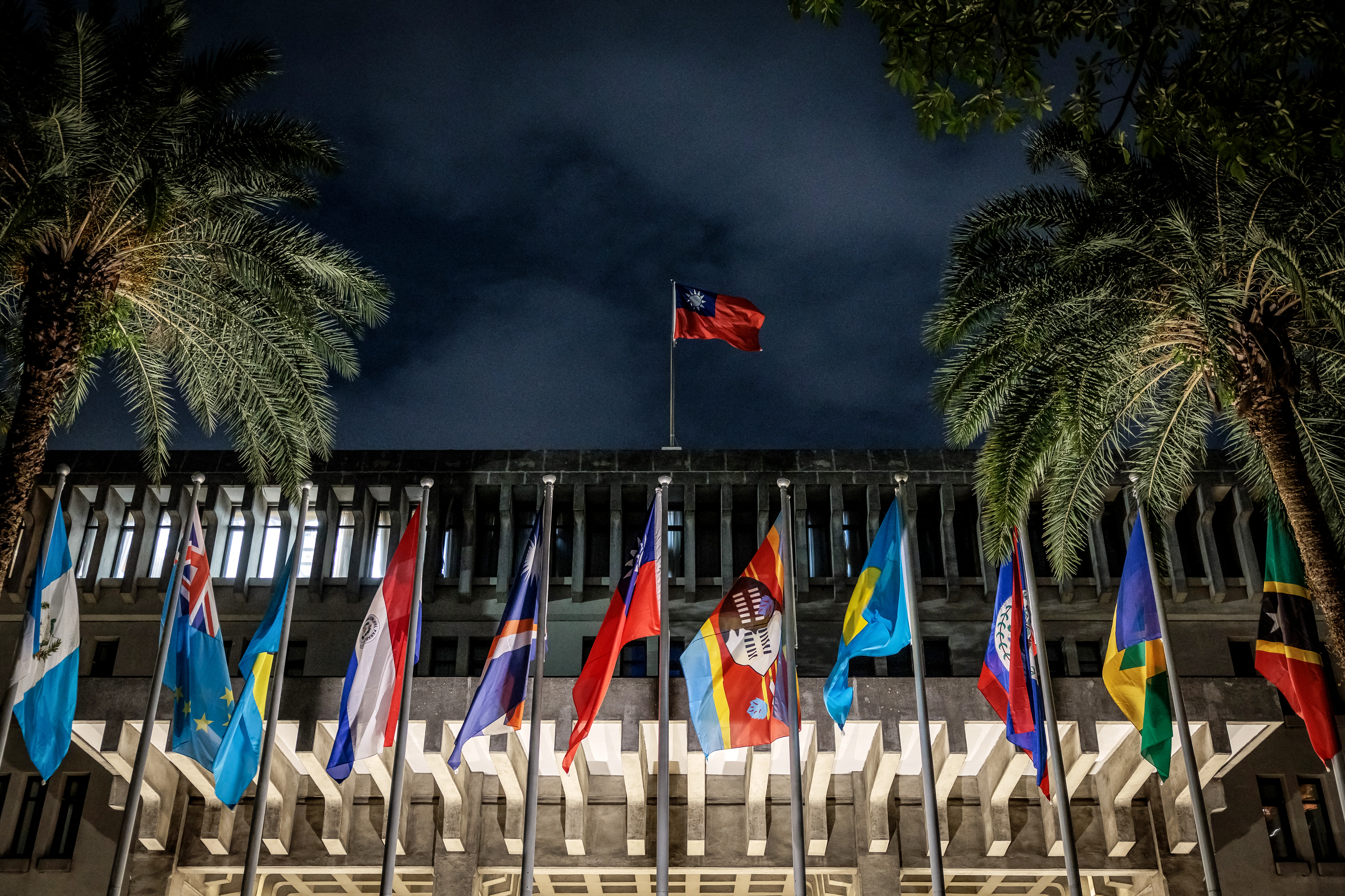Flags of Taiwan's remaining diplomatic allies on display outside the island's Ministry of Foreign Affairs. It's dark but the flags are illuminated. There is a Taiwan flag on top of the building. 