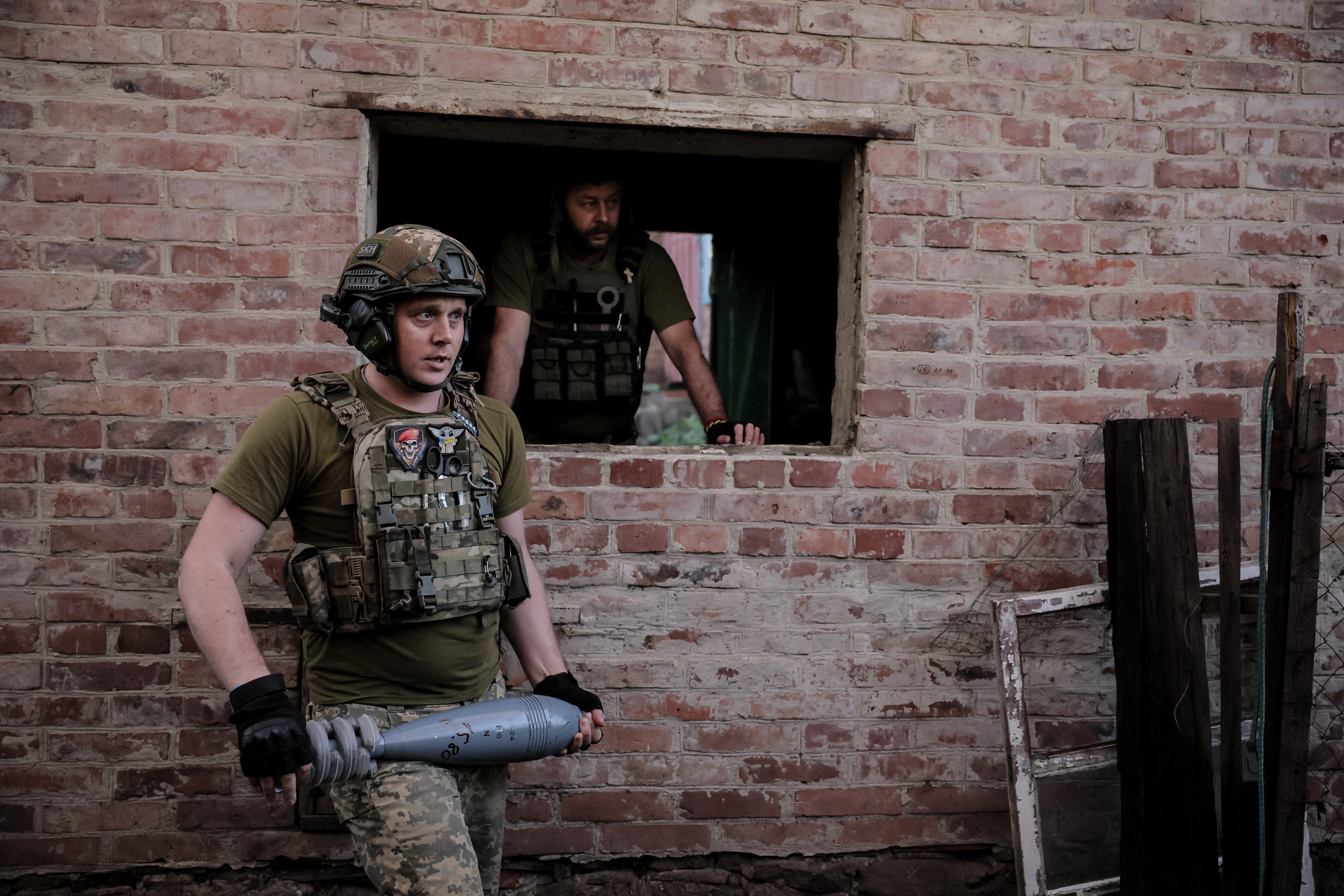 A Ukrainian soldier carrying a mortar, Another solider is looking out of a ruined building behind.