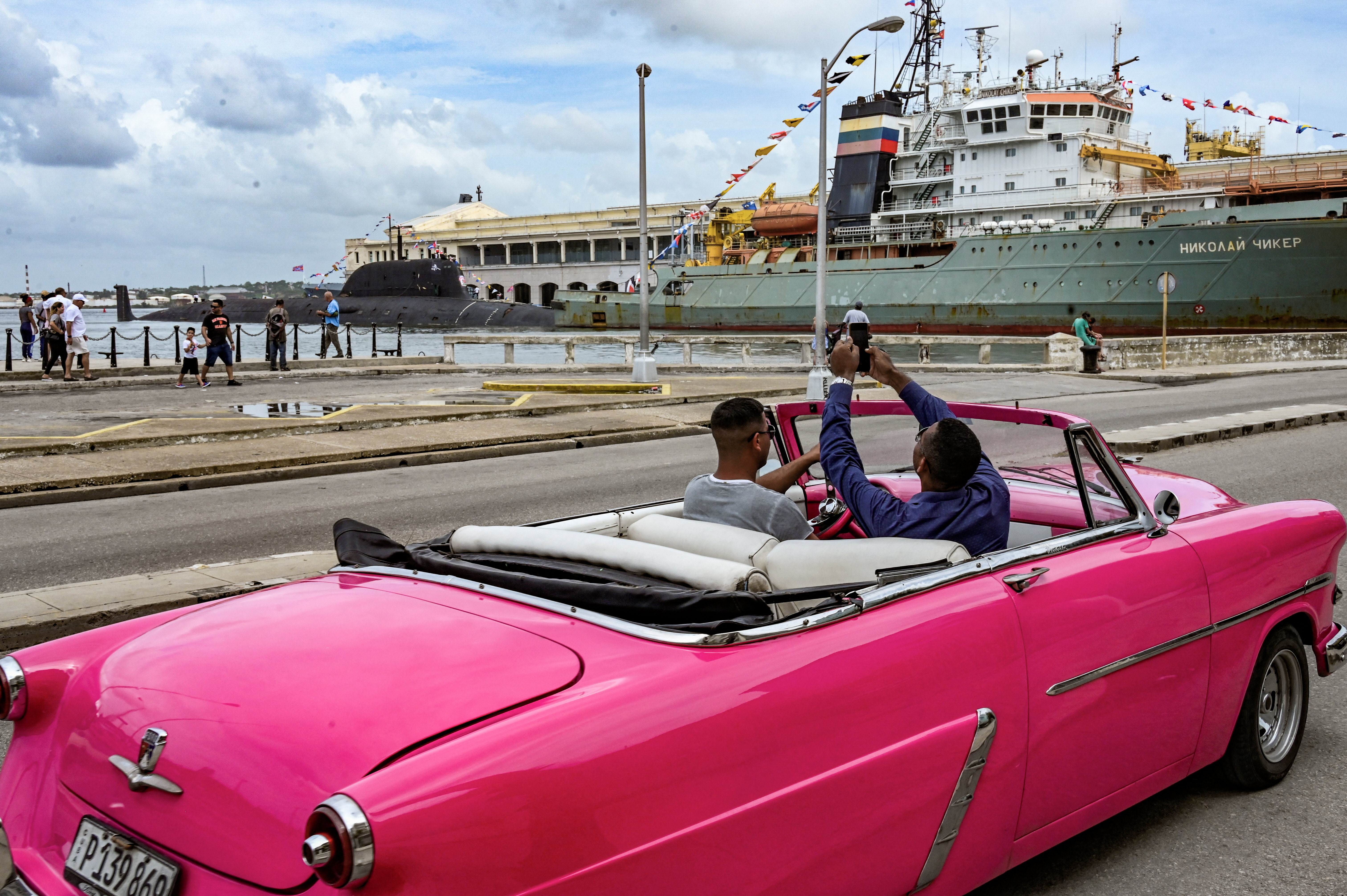A man taking pictures of the Kazan submarine and the Russian tugboat accompanying it from his classic open-topped car.