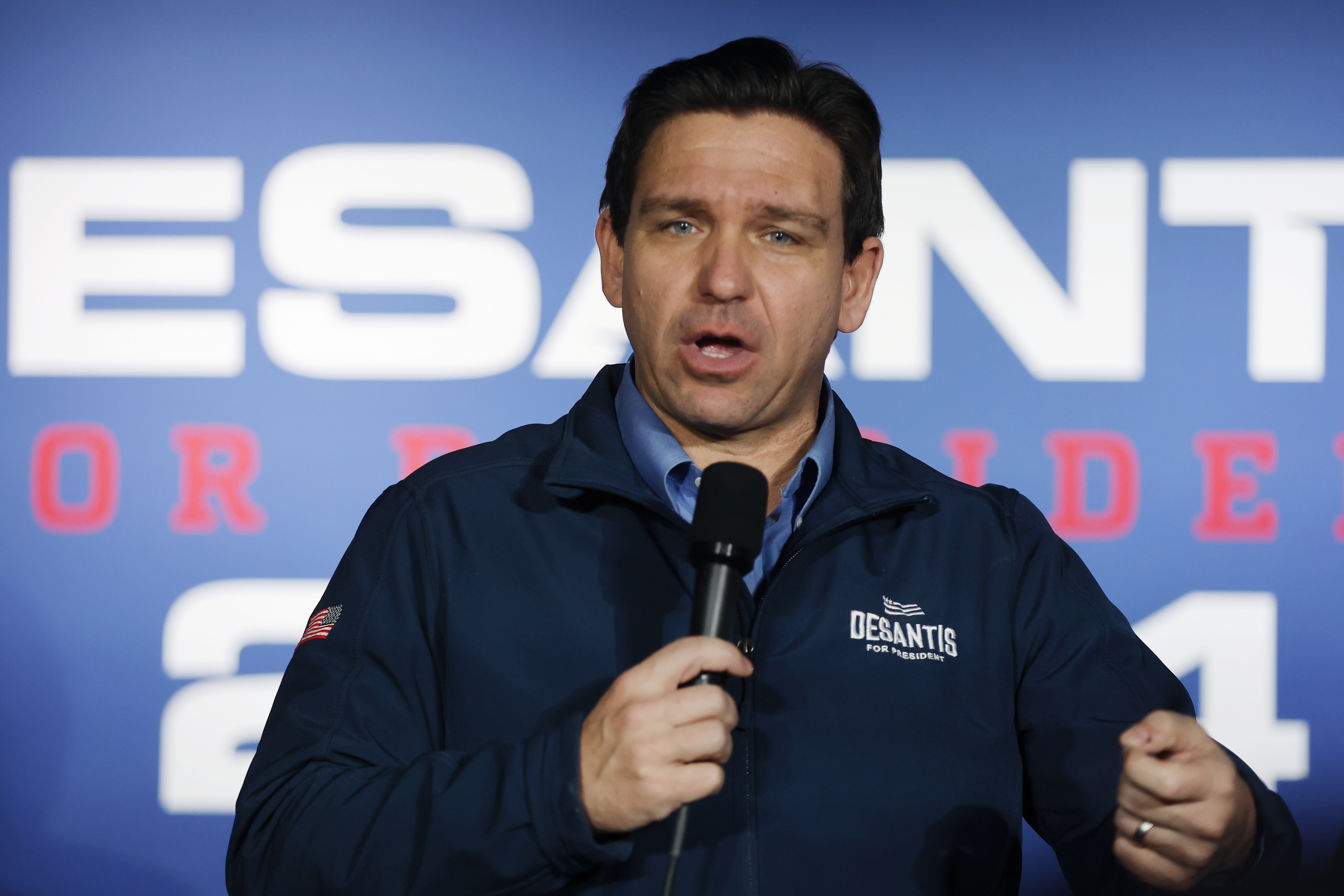 Governor Ron DeSantis speaks into a microphone in front of a screen that shows his presidential campaign logo.