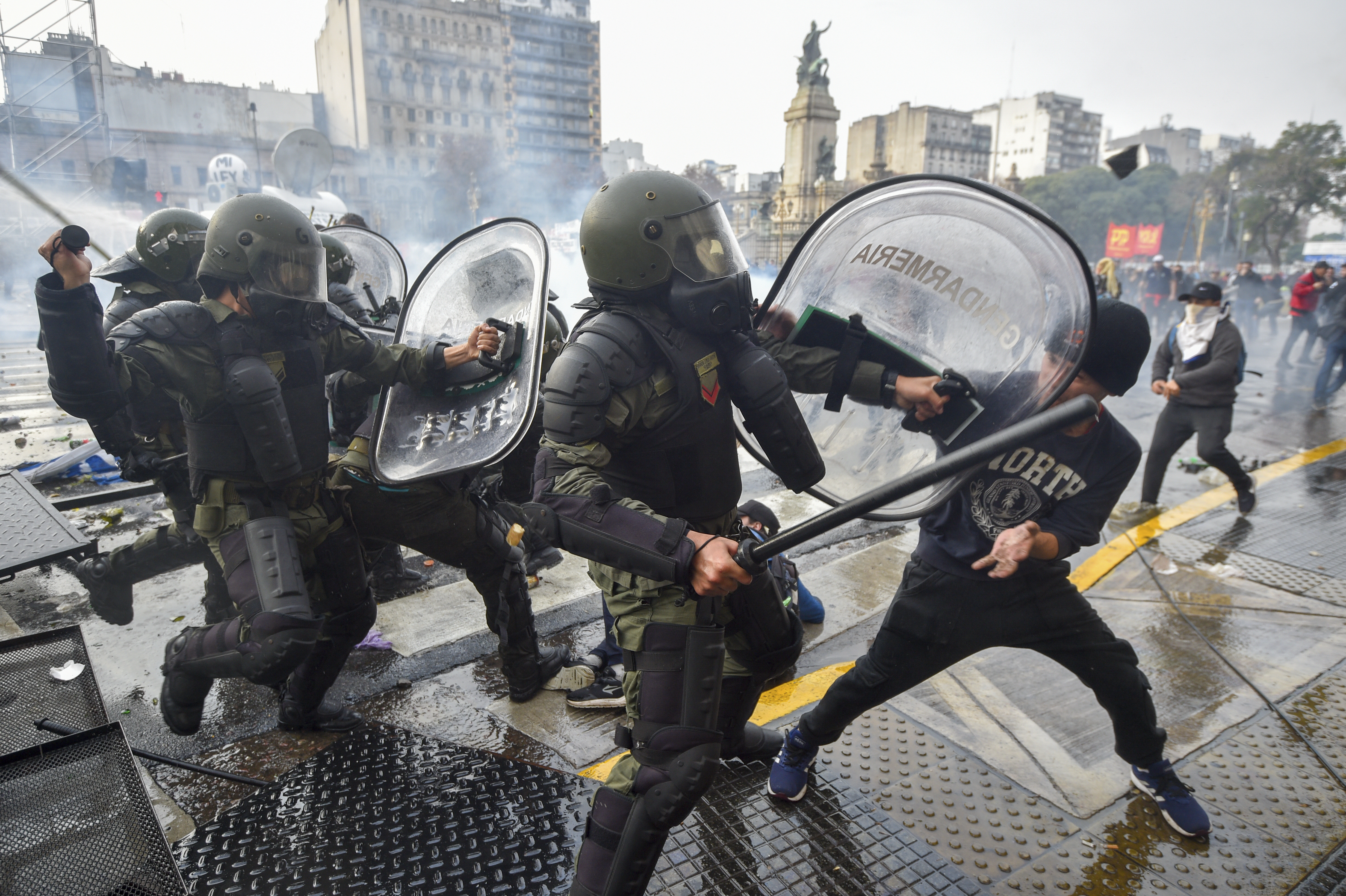 Police in riot gear and clear plastic shields clash with protesters in the street.
